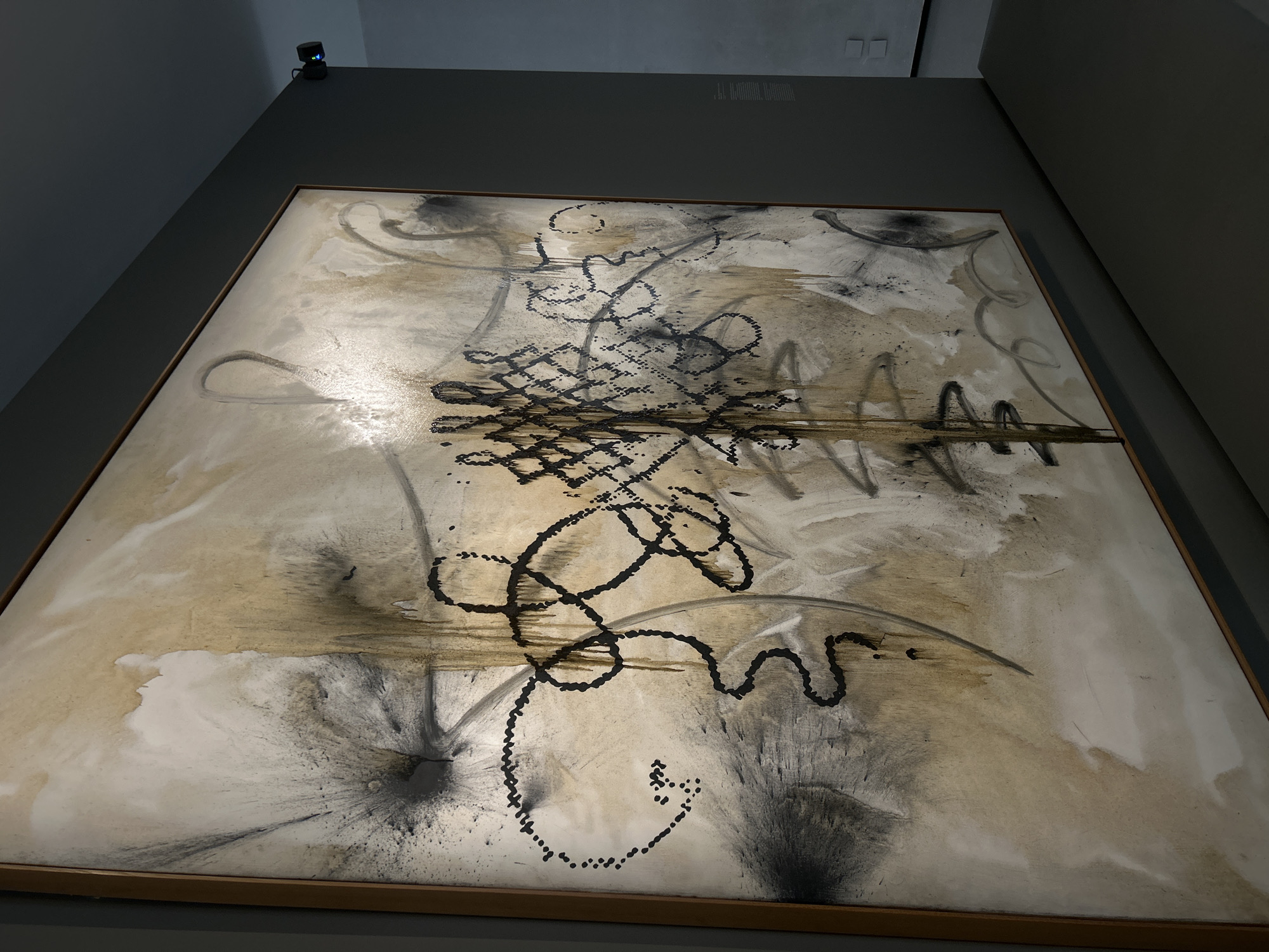 This is a medium shot, eye-level, angled photograph of a large abstract painting displayed in a gallery. The painting is predominantly abstract with a background of muted tones, including whites, creams, and light browns, with areas of darker, almost black, smudges and splatters. Overlaid on this background are thick, black, gestural brushstrokes and calligraphic marks. Some of these marks are fluid and curved, while others form more intricate, almost woven patterns, particularly in the center of the composition. There are also areas that resemble ink blots or spray paint effects, radiating outwards from dark centers. The painting is framed in a simple, light-colored wooden frame. The gallery setting is indicated by the dark gray walls and a subtle ceiling fixture visible in the top left corner, which appears to be a small security camera. The lighting is ambient and somewhat dim, creating soft reflections on the artwork. The overall impression of the artwork is one of dynamic energy and spontaneous expression, with a monochrome palette accented by bold black lines. There is no visible text within the image itself.
