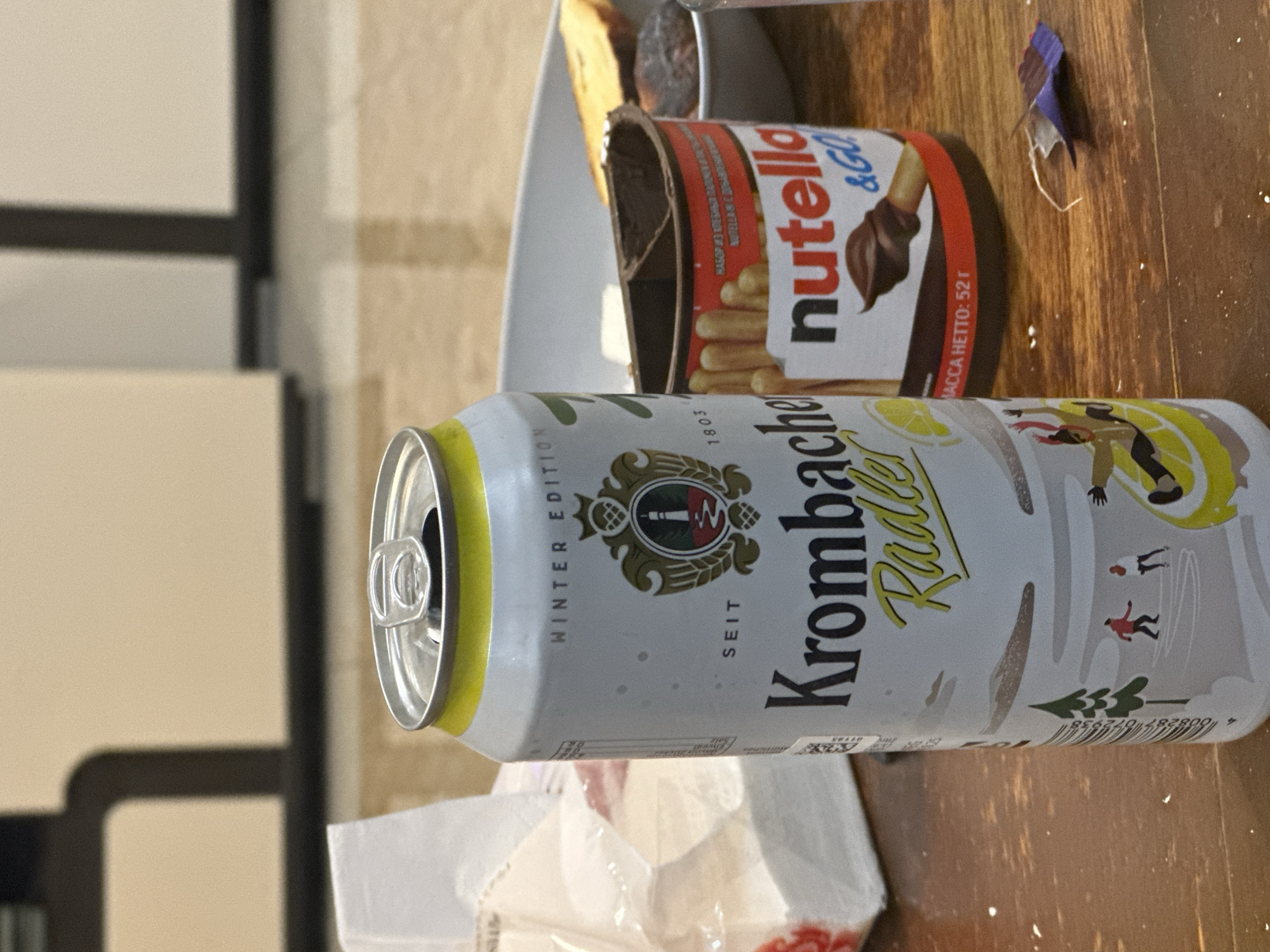 This image captures a close-up, rotated view (90 degrees clockwise) of a casual indoor setting featuring snacks and a beverage. The main subjects are an open Krombacher Radler can and an open Nutella & Go snack pack, both resting on a wooden surface, likely a table.
The Krombacher can, labeled "WINTER EDITION" and "Krombacher Radler," shows small illustrations of skiers, indicating a winter theme. Text on the can includes "SEIT 1803" and numerical codes like "56110" and "4 008287 072938." The can is open, with the pull tab visible.
Next to it is an open Nutella & Go pack, which appears to have been partially consumed. The branding "nutella &GO" is clearly visible, along with text in what appears to be Cyrillic script, "НАБОР ИЗ ХЛЕБНЫ... NUTELLA & C..." and "МАССА НЕТТО: 52г." This suggests it's a snack with breadsticks and a hazelnut spread.
In the background, a white bowl is partially visible, containing what looks like a piece of cake or pastry and another dark, irregular-shaped food item. A small, crumpled piece of blue and white paper or wrapper is also on the wooden surface near the Nutella pack. Below the Krombacher can, a crumpled white plastic bag or napkin is visible. The background further shows blurry light-colored walls or surfaces, indicating an indoor environment. The activity depicted suggests a relaxed moment, possibly a snack or refreshment break.