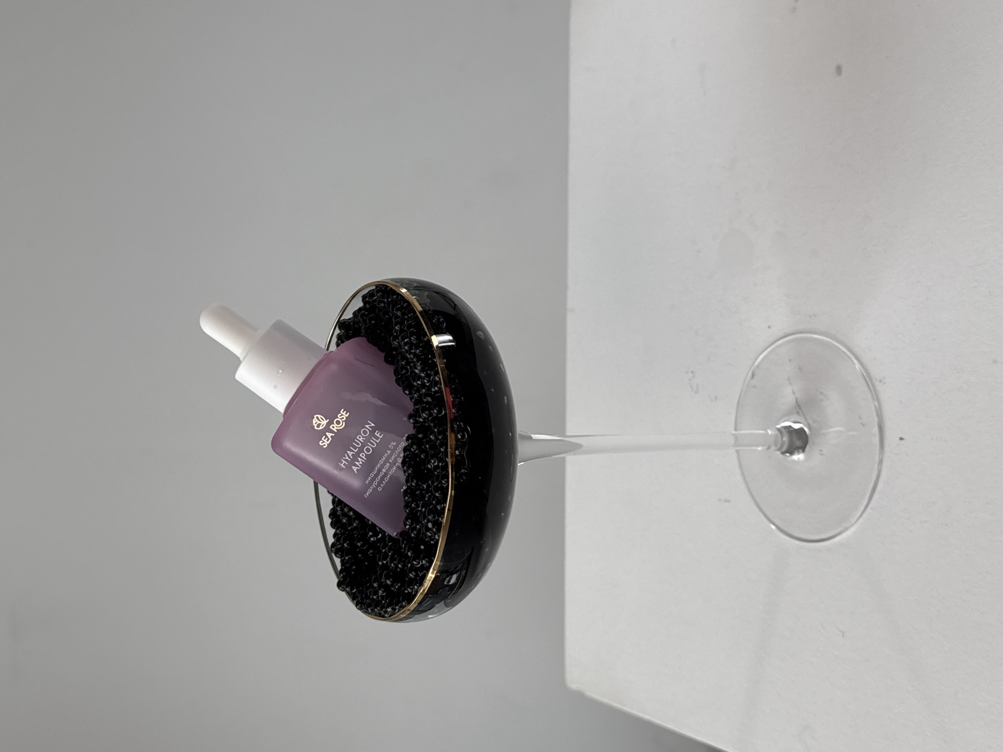 A sideways still-life photograph depicts a wine glass containing small black spheres, likely artificial caviar. Leaning against the caviar within the glass is a lavender bottle labeled "SEA ROSE HYALURON AMPOULE." The bottle has a white dropper top. The scene appears to be situated indoors, with a white wall visible to the right and a light grey wall to the left.
There is no indication of the location being Sochi, Russia.