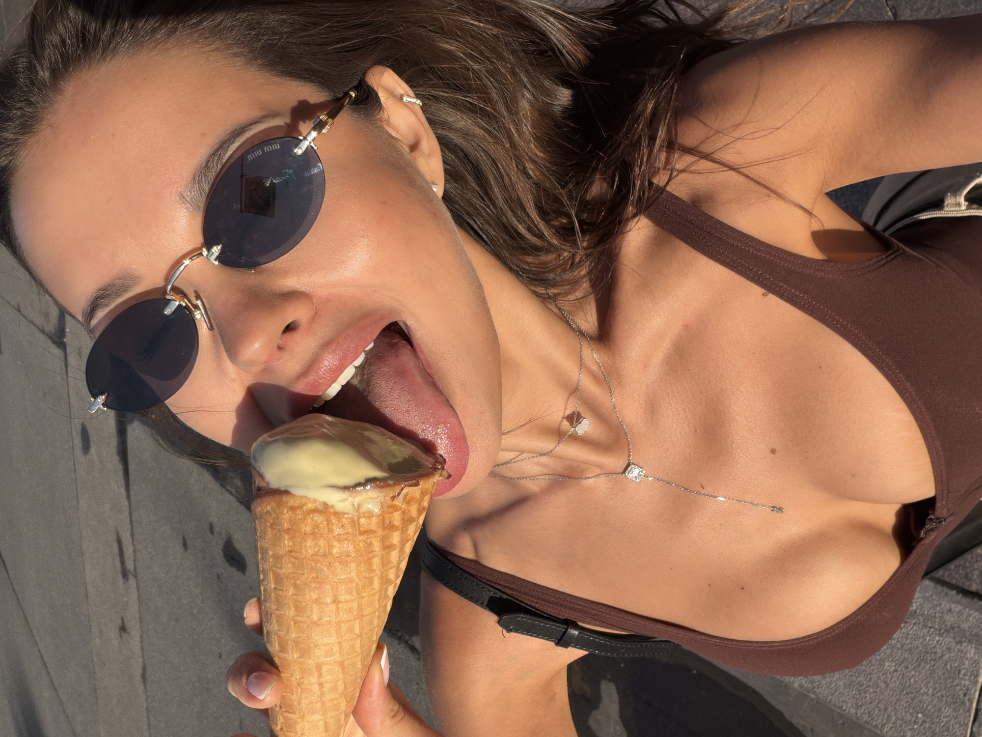 A woman, seen from the chest up, is outdoors, outdoors, in a city, with pavement and sunlight visible. She wears a brown tank top and two delicate silver necklaces. One necklace has a square pendant with a diamond, and the other has a smaller square pendant. She is wearing oval sunglasses with gold frames. The woman is holding an ice cream cone to her mouth, with her tongue extended to lick it. The ice cream appears to be vanilla-flavored with chocolate syrup. She has long, dark brown hair. The lighting suggests it is daytime, likely sunny. The scene is not specifically identifiable as Saint Petersburg, Russia, based on visual cues alone.