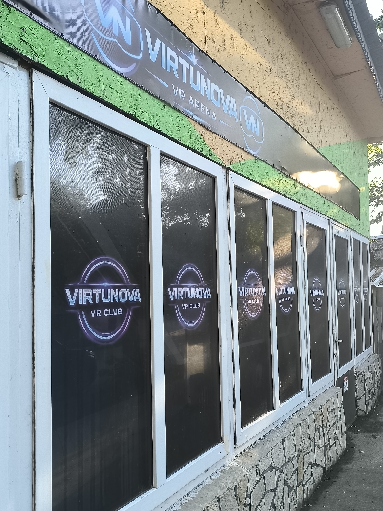 The image captures the exterior facade of a commercial building, likely a VR arcade or club, in Trastenik, Bulgaria. The building features a series of white-framed windows, seven visible in total, extending along its length. These windows are covered with dark, reflective material, each displaying a circular logo and the text "VIRTUNOVA VR CLUB." The reflection of trees or foliage can be subtly seen in the upper parts of the window coverings.
Above the windows, a green-painted wooden frieze is visible, topped by a large black banner. This banner prominently displays "VIRTUNOVA VR ARENA" in blue and white lettering, accompanied by a stylized "VN" logo. The wall above the banner appears to be made of light-colored wood panelling or OSB board, possibly painted, and has a single fluorescent light fixture attached to it. The upper part of this wall shows warm, golden light, suggesting it's daytime, possibly late afternoon or early morning with sunlight hitting the building.
Below the windows, the foundation of the building is clad with rough, grey and beige stone blocks, giving it a somewhat rustic appearance. The ground in front of the building is a paved or concrete surface. The overall impression is that of a functional, albeit modestly constructed, commercial establishment. No people or specific activities are depicted in the static shot.