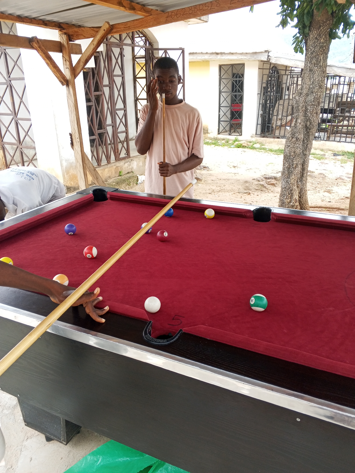 A teenage African male stands behind a red felt pool table, holding a pool cue and looking down at the table. He is wearing a pale pink t-shirt. The pool table has several balls on it, including a purple ball, a red and white striped ball labeled "1", a red ball labeled "1", a green and white striped ball labeled "14", a white ball, a yellow ball, and a blue ball. A hand is visible on the left side of the frame, reaching towards the pool table. The pool table is located outdoors, under a wooden and metal roof structure. In the background, there is a building with barred windows and a tree. The ground appears to be dirt and gravel. A green plastic sheet is visible on the ground in the lower right corner. The scene is likely located in Lokoja, Nigeria.