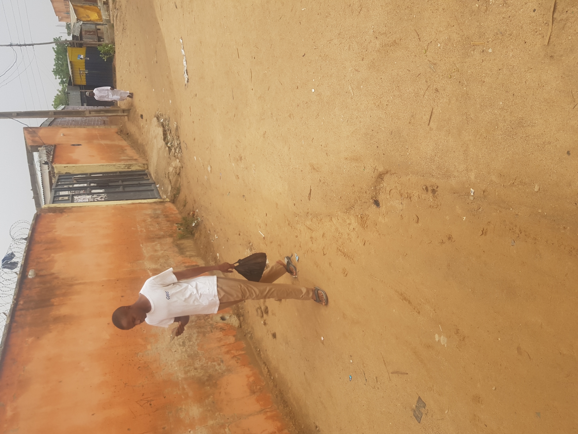 The scene captures a street in Dabawa, Nigeria. A young man, wearing a white t-shirt and khaki pants, walks on a dirt road. He carries a dark-colored bag. To the left, a wall is topped with barbed wire. A man in white is seen walking in the distance. The environment includes basic infrastructure like buildings and power lines, set against the backdrop of a dusty, unpaved road. The overall setting suggests a developing urban area.
