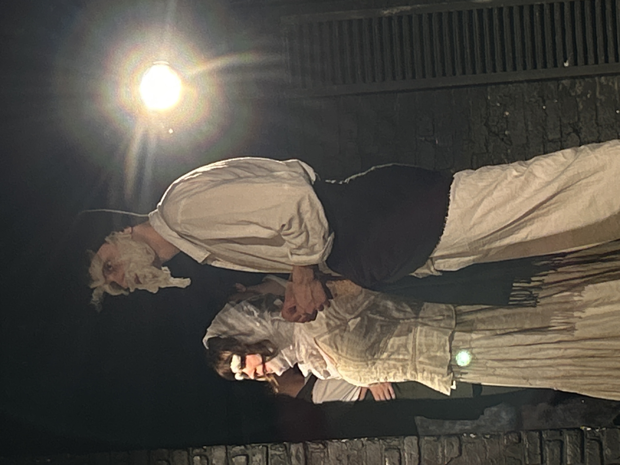 Two actors are on stage in a dark, unidentified city in Unknown. An older actor with a white beard, wearing a white shirt, black vest, and white pants, is holding hands with a younger actress who is wearing a long white dress and white head covering. The actors are standing on a raised platform resembling a stone wall. Above them is a grate. A light shines brightly on the stage.