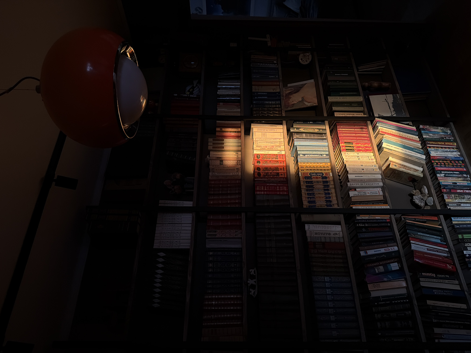 The image shows a dimly lit bookshelf filled with stacks of books. A spherical lamp with an orange exterior and a white interior is positioned on the left side of the frame. The light from the lamp casts a warm glow on some of the books, highlighting their spines and titles. The bookshelf is organized into several sections, with books arranged vertically in each section. The books vary in size, color, and thickness. The light emphasizes the textures and details of the book covers, creating a play of light and shadow across the shelf. Some titles are visible, featuring Russian words and names. The location is Khimki, Russia.