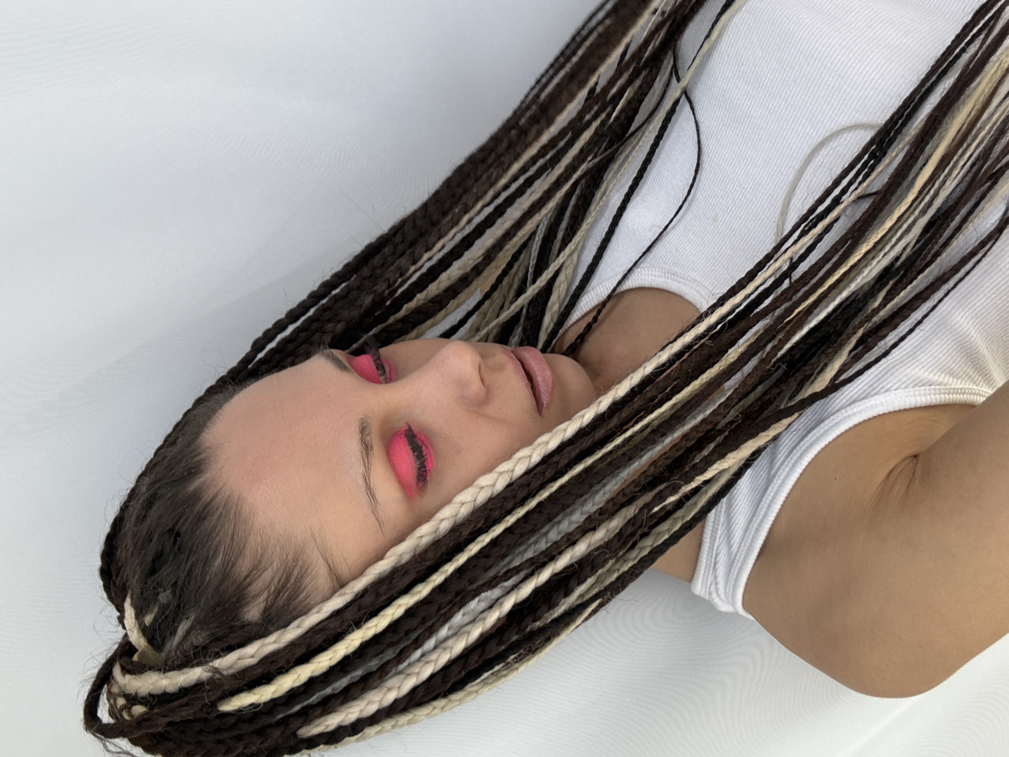 A person lies on a white surface. They wear a white tank top and have brown and blonde braided hair. Their eyelids are painted with bright pink eyeshadow. The location is unspecified but could be Rusanovo, Russia.