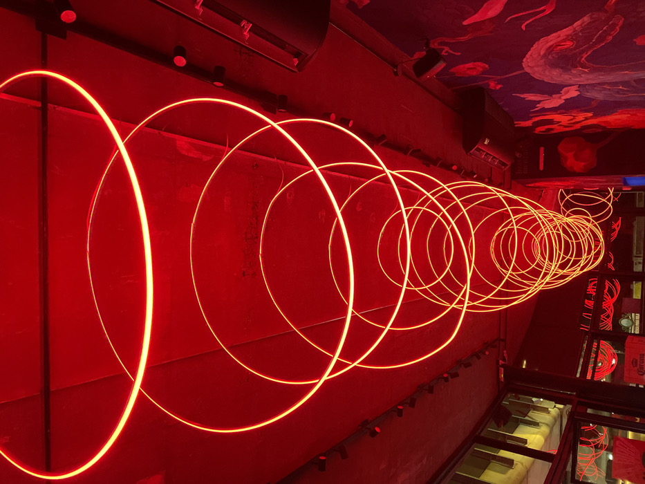 A line of glowing red circles ascends in diminishing size against a red wall. The circles, illuminated with neon or LED lights, create a tunnel-like illusion. The background includes a patterned ceiling with dragon motifs, dark-colored track lighting, and a glimpse of a lower level with a glass floor. Reflections on the glass reveal fragments of the circular light design.