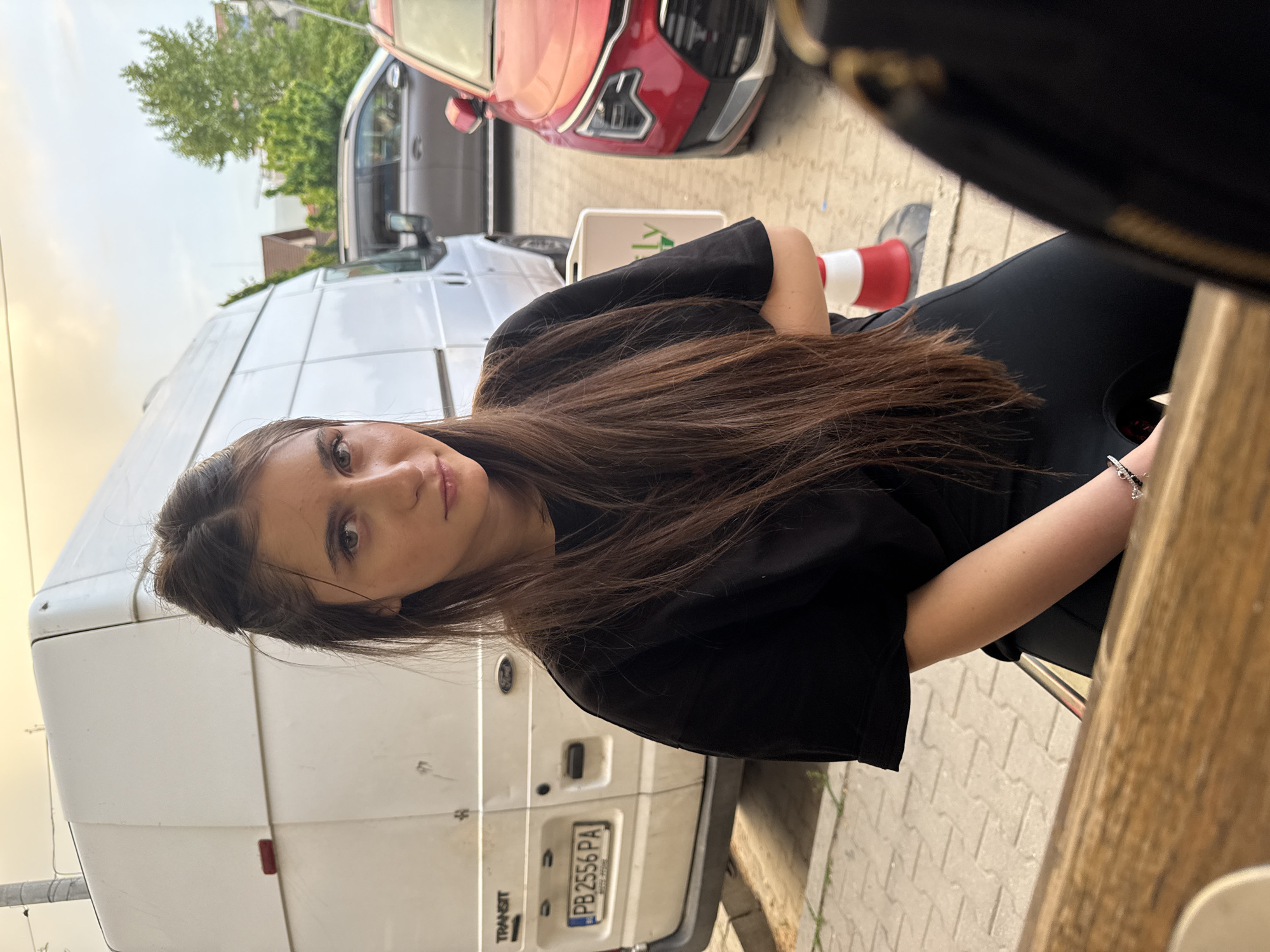 A young woman with long brown hair is seated outdoors at a table. She is wearing a black top. Behind her is a white van with a license plate reading "PB 2556 PA." A red car is parked nearby. A red and white traffic cone sits on the sidewalk. The location is possibly Plovdiv, Bulgaria. The woman appears to be looking at the camera. The image is taken at an angle, slightly rotated.