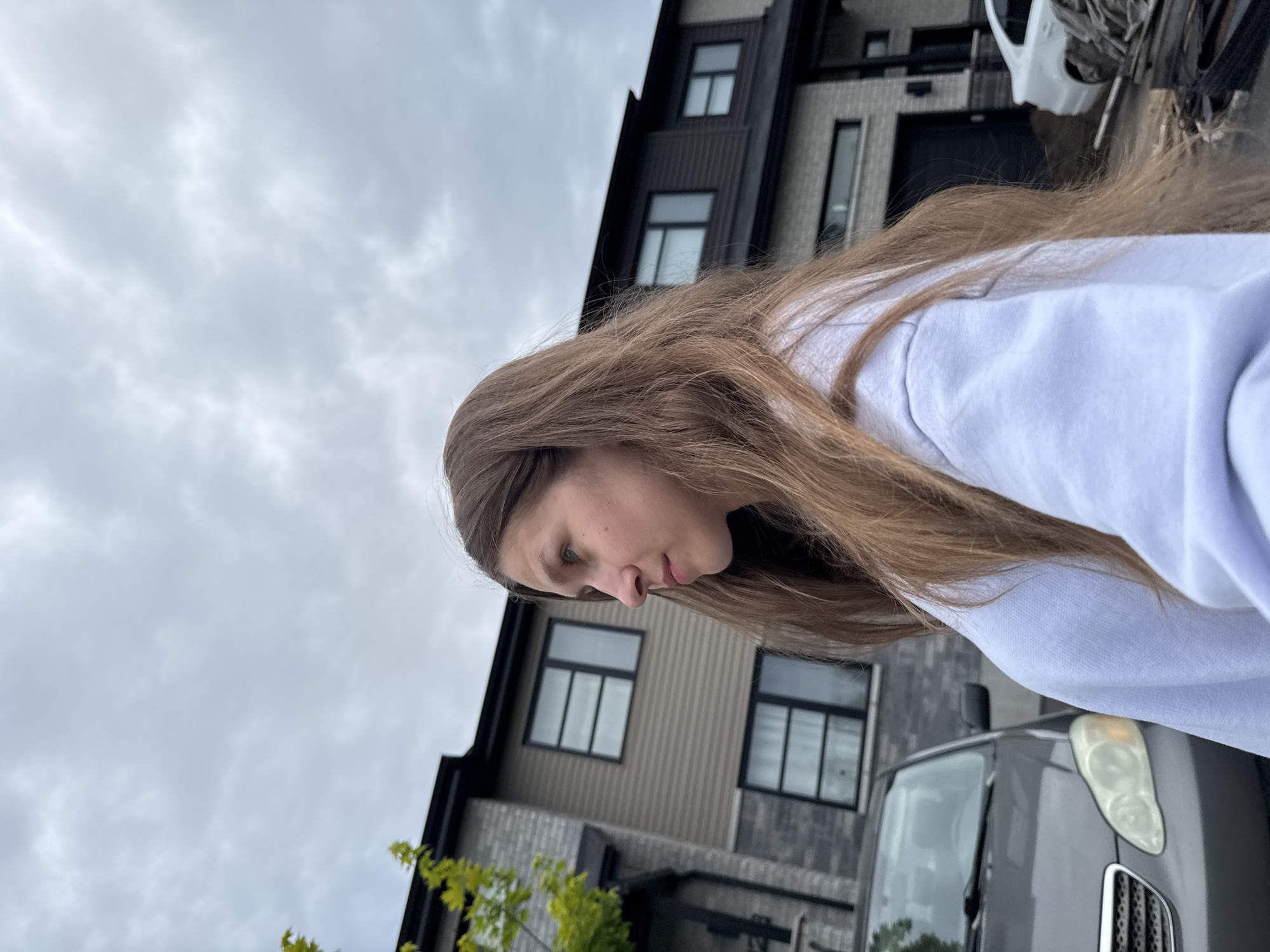 A young woman with long, brown hair is shown in profile, looking to the left. She is wearing a light-colored sweatshirt. In the background, a two-story house with grey brick and horizontal siding is visible. The house has multiple windows with dark frames. To the left of the house, a portion of a gray car is visible. The scene appears to be outdoors in a residential area, possibly in Gatineau, Canada, given the architectural style and the overcast sky.