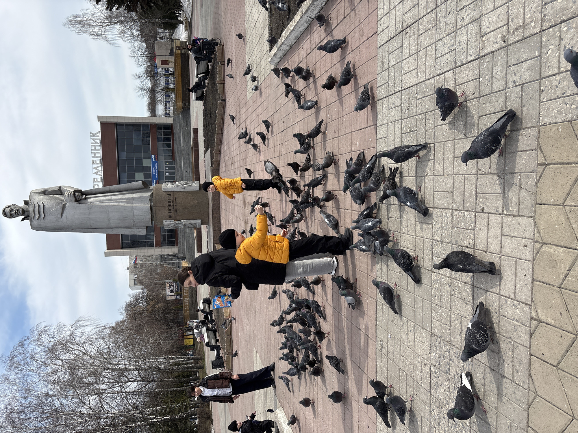 A medium shot shows a city square with people and many pigeons. Two people are standing, feeding the birds. One person is wearing a yellow jacket and black hat. The other person is wearing a black jacket and hair tied up. A group of people are standing to the left. A statue and building with Cyrillic lettering are in the background. The building has the Russian flag displayed. The ground is paved with tan and red bricks. The location is likely Ulyanovsk, Russia based on the cyrillic lettering.