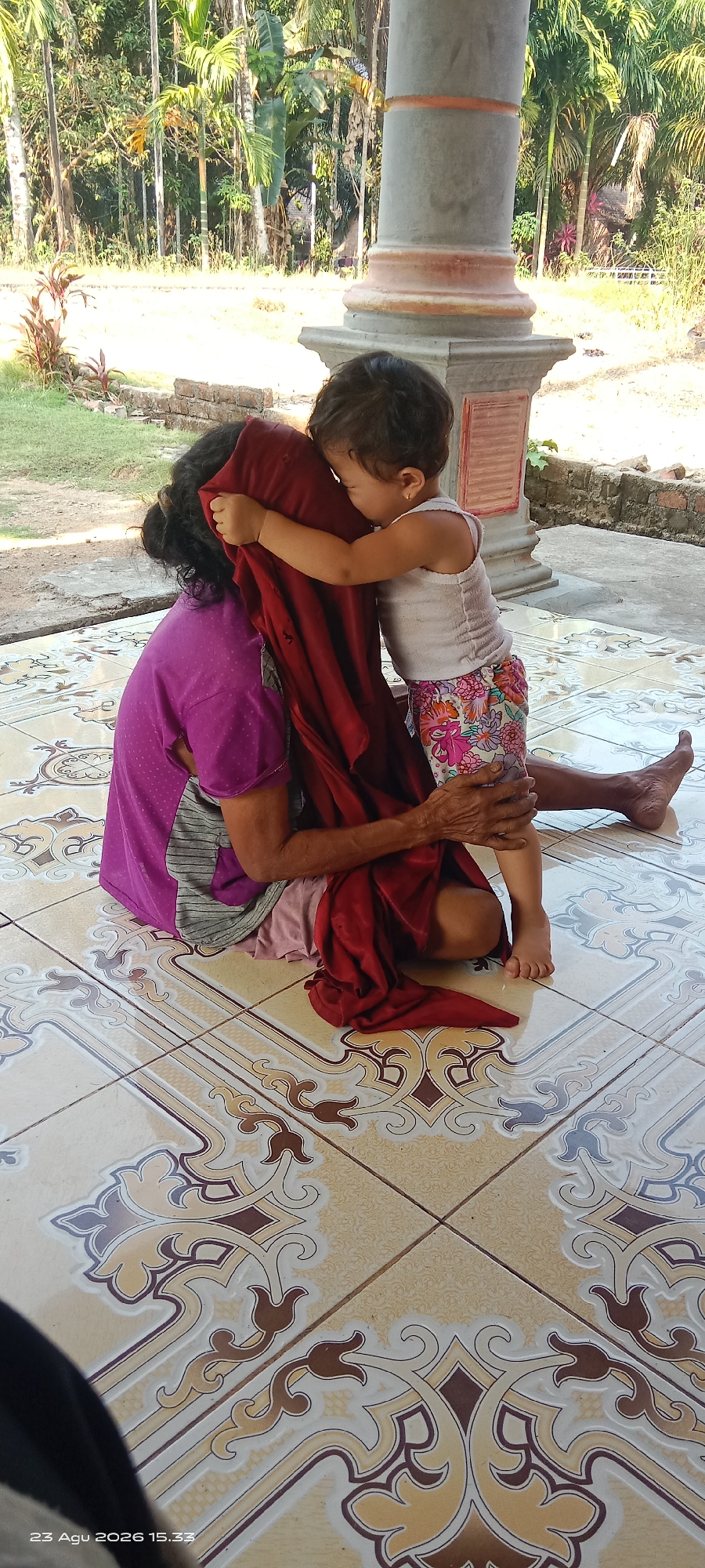 This is a full-frame, eye-level, vertical shot of a young child embracing an adult from behind, both seated on a tiled floor. The adult, wearing a purple shirt and gray and white striped fabric, is partially obscured by a dark red cloth. The child, wearing a white tank top and floral patterned shorts, has their arms wrapped around the adult's neck, their face hidden by the red cloth. The child's right leg is visible, bent at the knee. The adult's legs are extended, with their right foot visible on the floor. The tiled floor features an ornate pattern of beige, brown, and off-white. In the background, out of focus, are lush green trees and foliage, suggesting an outdoor or semi-outdoor setting. A stone pillar with orange banding is to the right of the subjects. The bottom of the frame displays a timestamp: "23 Agu 2026 15.33". The image appears to have been taken in Jajaran Baru, Indonesia.