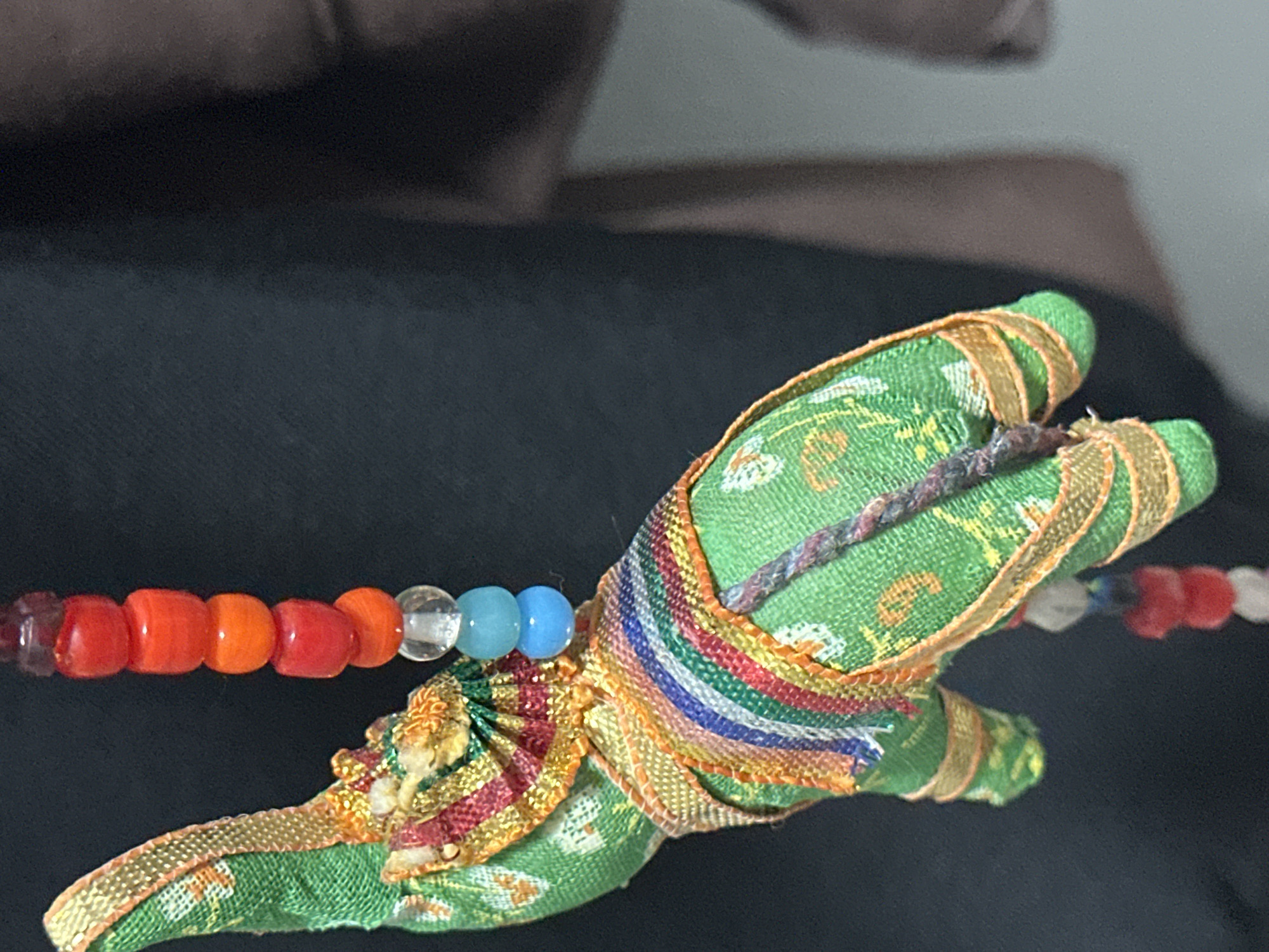 The image shows a close-up of a green fabric elephant figurine with colorful decorative trim, suspended by a beaded string. The elephant has a patterned fabric exterior and appears to be handmade. The beaded string consists of red, orange, clear, and blue beads. The background is dark, suggesting an indoor setting. There is no identifiable location within the city of Nevinnomyssk, Russia.