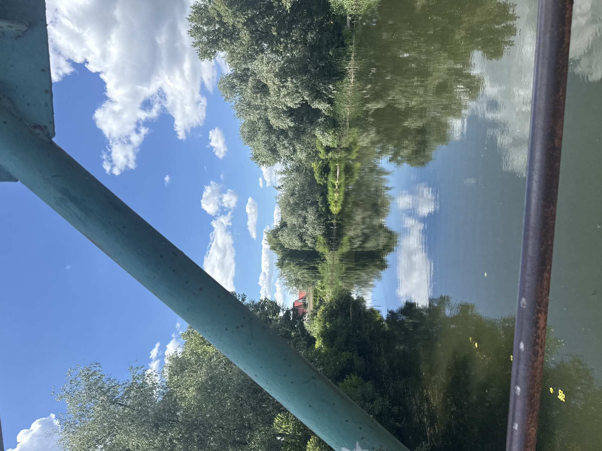 This is a photograph taken from a bridge overlooking a body of water, likely a river or canal, in Obukhivka, Ukraine. The water is calm and reflects the sky, trees, and clouds. Lush green trees line the banks of the water, with some reeds and vegetation visible. In the background, a few houses with red roofs can be seen nestled amongst the trees. The sky is a bright blue with scattered white, fluffy clouds. A teal-colored railing is prominently visible in the foreground, diagonally crossing the frame, suggesting the vantage point is from a bridge or similar structure. The overall scene depicts a tranquil natural environment within a populated area.
