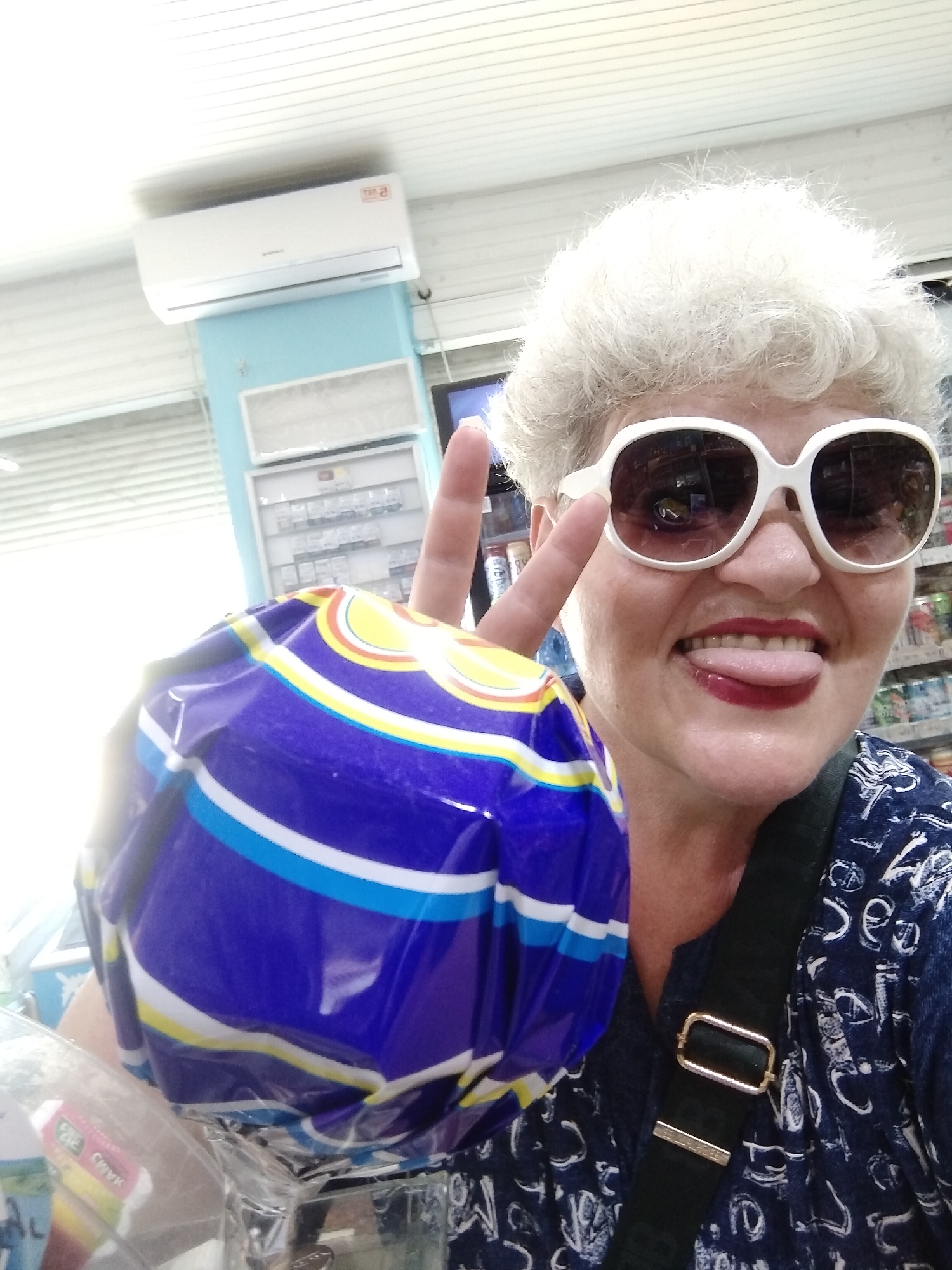 The image shows a woman with grey curly hair, large white sunglasses, and red lipstick. She is sticking her tongue out and making a peace sign with her right hand. She is holding a blue, white, and yellow striped object, possibly a bag of chips, in her left hand. She is wearing a dark blue top with a white pattern. A dark green strap with a gold buckle is visible across her shoulder. In the background, there is a white air conditioner unit mounted on a light blue wall. Shelves with various products are visible behind her. The overall setting appears to be a store or market. There are no discernible elements that specifically indicate the location within Horishni Plavni, Ukraine.