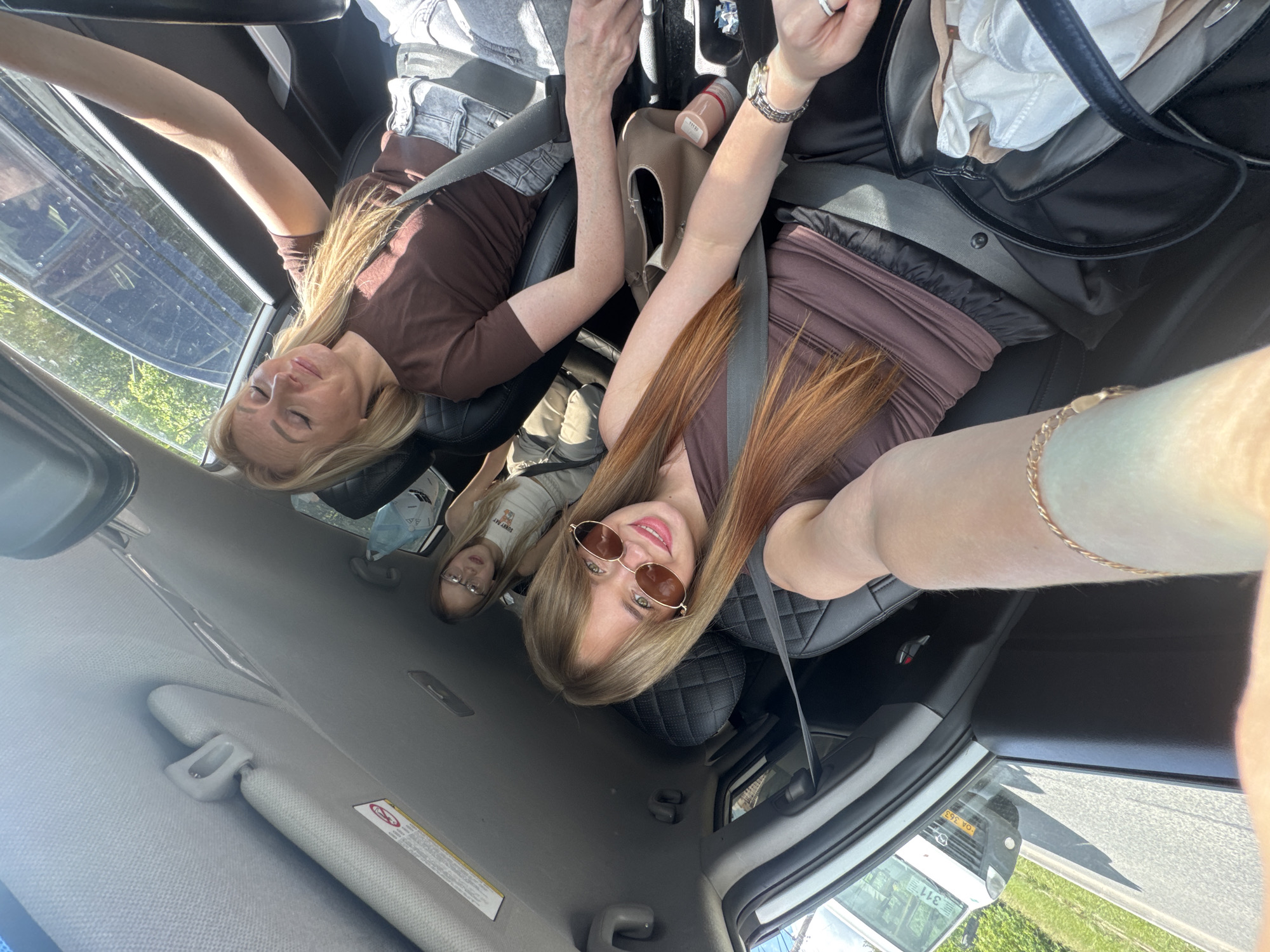 Three women are in a car. Two women in the front seats are wearing seatbelts and brown clothing. One woman is wearing sunglasses. A younger woman is in the back seat. The location may be Izhevsk, Russia.