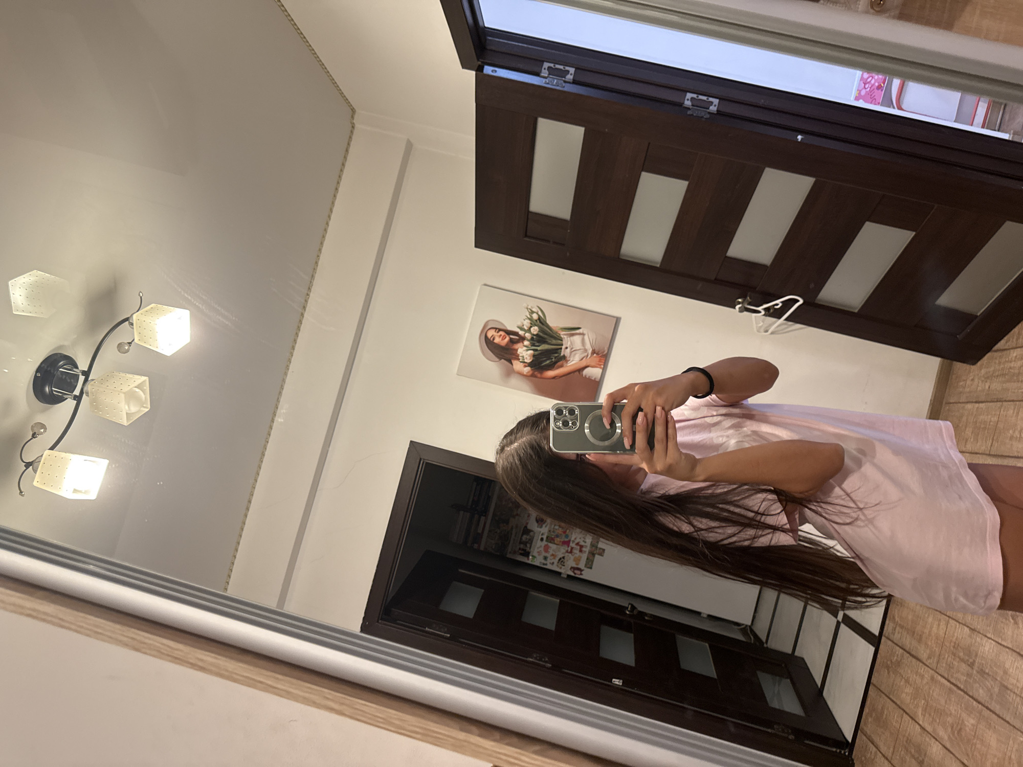 The image shows a young woman with long brown hair taking a mirror selfie in a room. She is wearing a pale pink t-shirt. In the background, there are two closed dark wooden doors with frosted glass panes, a painting on the wall of a woman holding tulips, and a light fixture on the ceiling. The floor is wooden. The reflection in the mirror also shows a glimpse of what appears to be a hallway or another room. There is no identifiable information that places this scene in the city of Chișinău, Moldova.