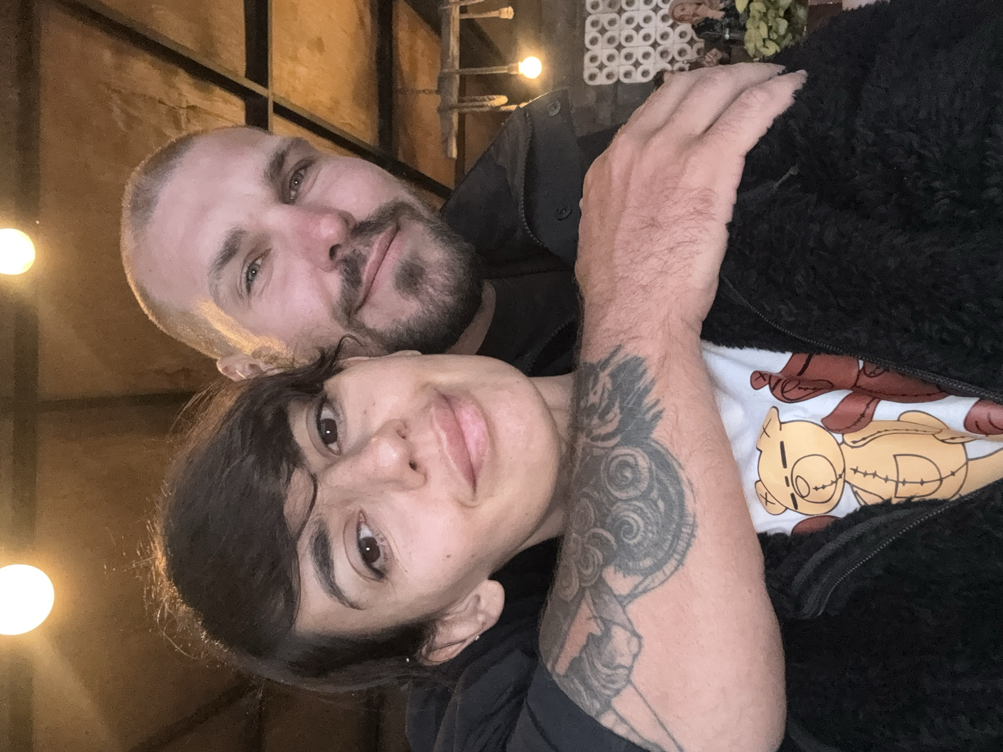 A close-up, selfie-style photograph features a man and a woman embracing indoors. The woman, on the left, has dark hair and is wearing a black jacket over a white t-shirt with a cartoon teddy bear print. Her left arm is visible, adorned with tattoos. The man, on the right, has a shaved head and a beard, and is wearing a dark jacket. He has his arm around the woman. The background suggests an indoor setting with a wooden ceiling and ambient lighting from overhead bulbs. The environment does not resemble Kintamani, Indonesia.