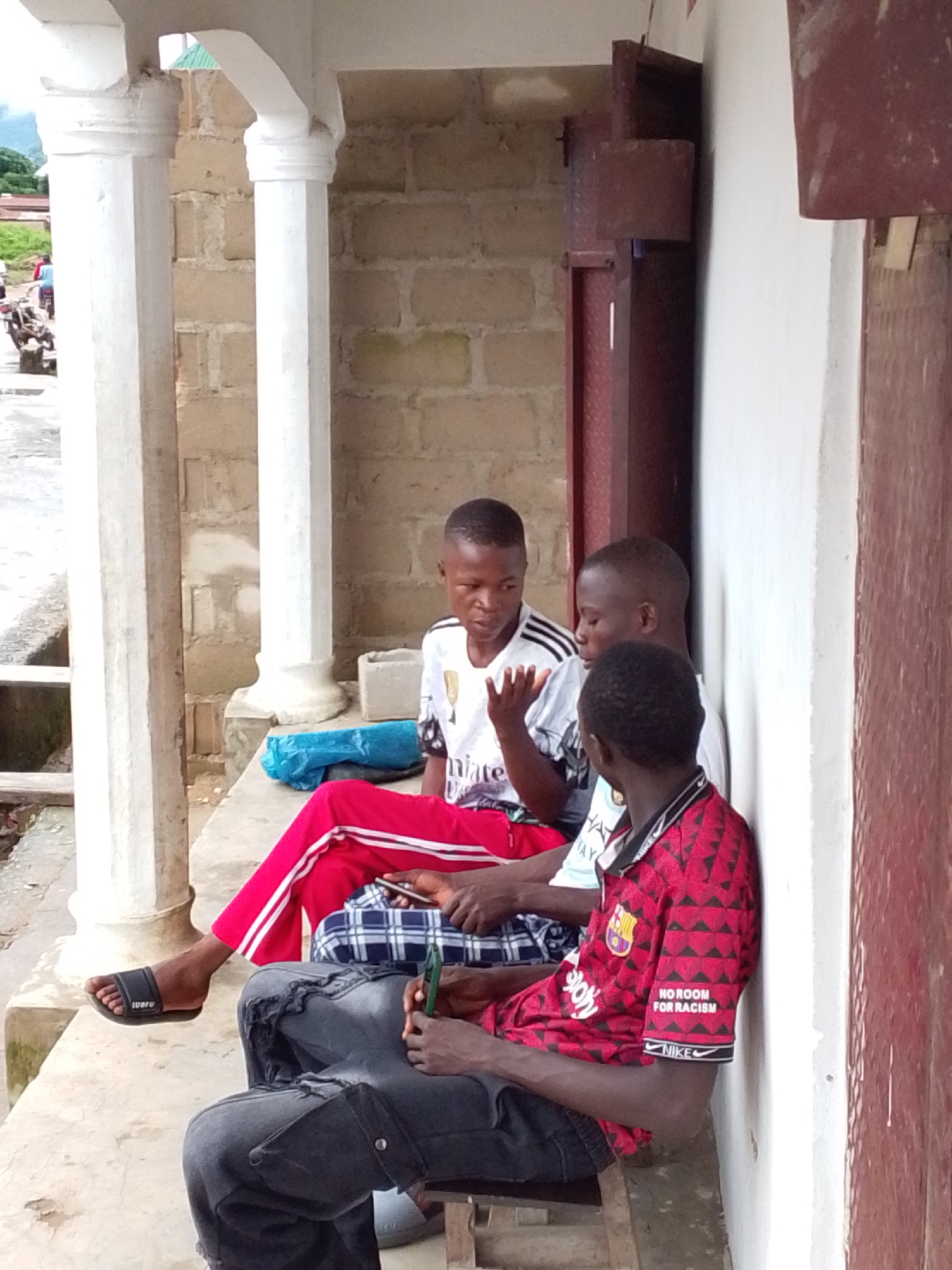 Three young males are sitting on a concrete ledge in front of a building with white columns and a brick wall. One male on the left wears a white t-shirt with black stripes on the sleeves and red and white striped pants. He is looking at his phone. The second male, in the middle, wears a white t-shirt and is looking at the first male. The third male on the right wears a red and black patterned shirt with the text "NO ROOM FOR RACISM" on the sleeve. He is looking down at his phone. A blue plastic bag and a checkered fabric are on the ledge. A motorcycle is visible in the background to the left. The scene appears to be in the city of Serti, Nigeria.