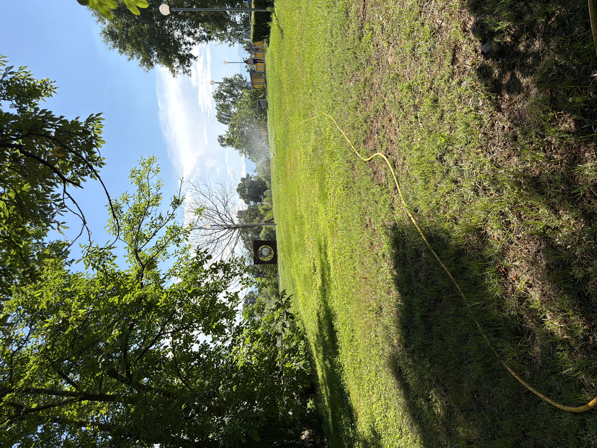 The image depicts a sunny outdoor scene in Pazardzhik, Bulgaria, likely within a park or recreational area. A bright yellow garden hose snakes across a lush green lawn, leading towards a sprinkler that is actively watering the grass. In the background, two individuals are walking on a paved path behind a yellow fence. The environment features mature trees, some with dense foliage and others bare, casting shadows on the ground. A unique circular sculpture is visible on a grassy mound in the mid-ground. The sky is a clear blue with scattered white clouds, suggesting fair weather. The overall impression is one of a well-maintained public space on a pleasant day.