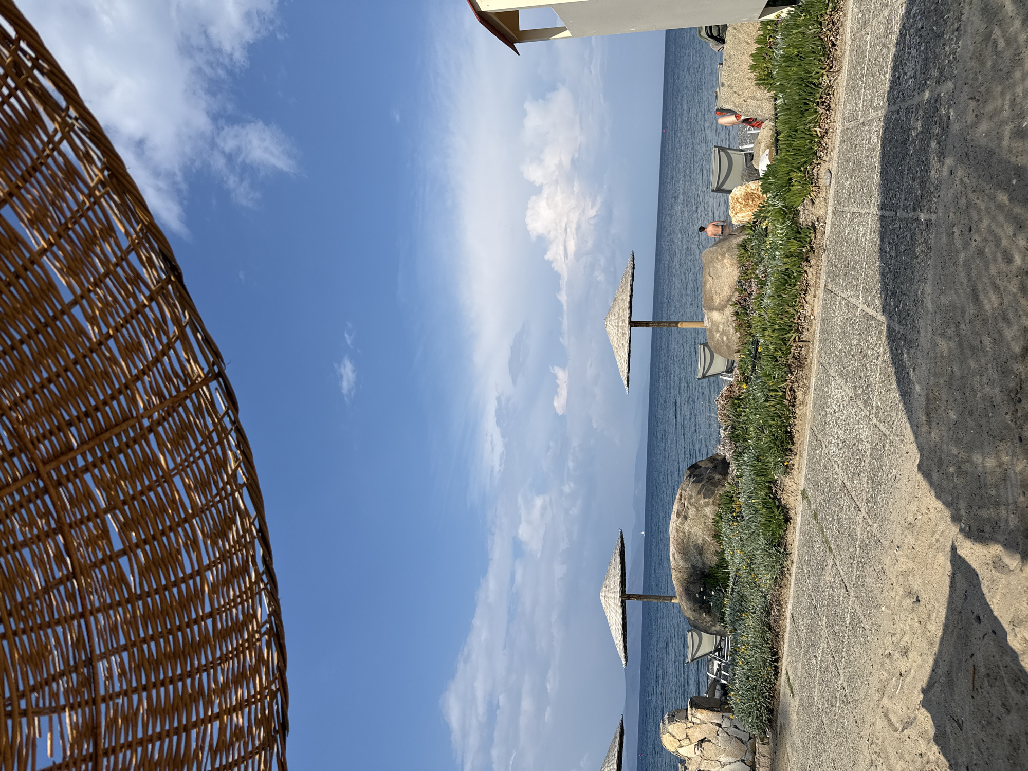 The image captures a sunny day at a coastal location in Kos, Greece. From left to right: Part of a woven beach umbrella is visible. A stone path runs along the edge of a raised area with greenery, leading towards the sea. Wicker umbrellas dot the shoreline, and chairs are visible on the beach below. Two individuals are visible in swimsuits on the beach. The sky is blue with scattered clouds, reflecting on the water. In the upper right corner is a small building.
