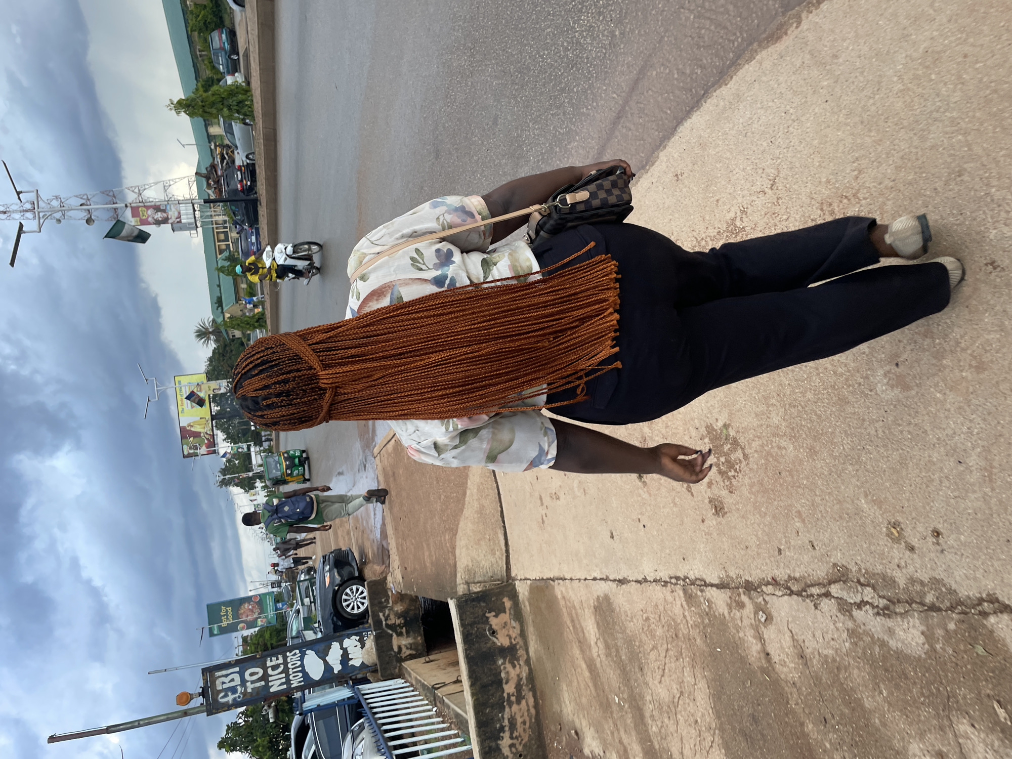 A full-shot, over-the-shoulder rear view of a woman walking on a sidewalk in Jos, Nigeria. The woman has long, braided auburn hair and is wearing a light-colored floral shirt and dark pants. She carries a checkered shoulder bag. In the background, traffic moves along a street under a cloudy sky. A man with a backpack walks away from the camera on the sidewalk, and a person rides a motorcycle on the street. Billboards and buildings line the street, with a cell tower visible in the distance. The sidewalk is made of concrete, and a drainage ditch runs alongside it. The overall scene suggests a busy urban environment.