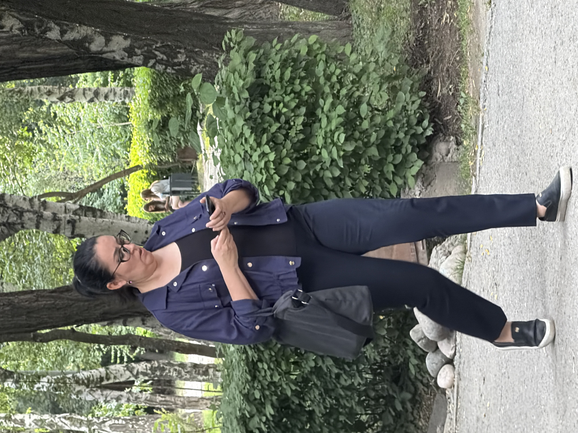 A woman is walking outdoors, looking at her phone. She has dark hair pulled back into a ponytail, wears glasses, a dark blue jacket over a black top, and dark pants. A black bag is slung over her left shoulder. She is wearing black slip-on shoes. She is walking on a paved path with a dense green hedge to her right and trees behind her. A pile of smooth stones is visible to her left. In the background, two other people are seated near a low wall. The environment appears to be a park or garden.