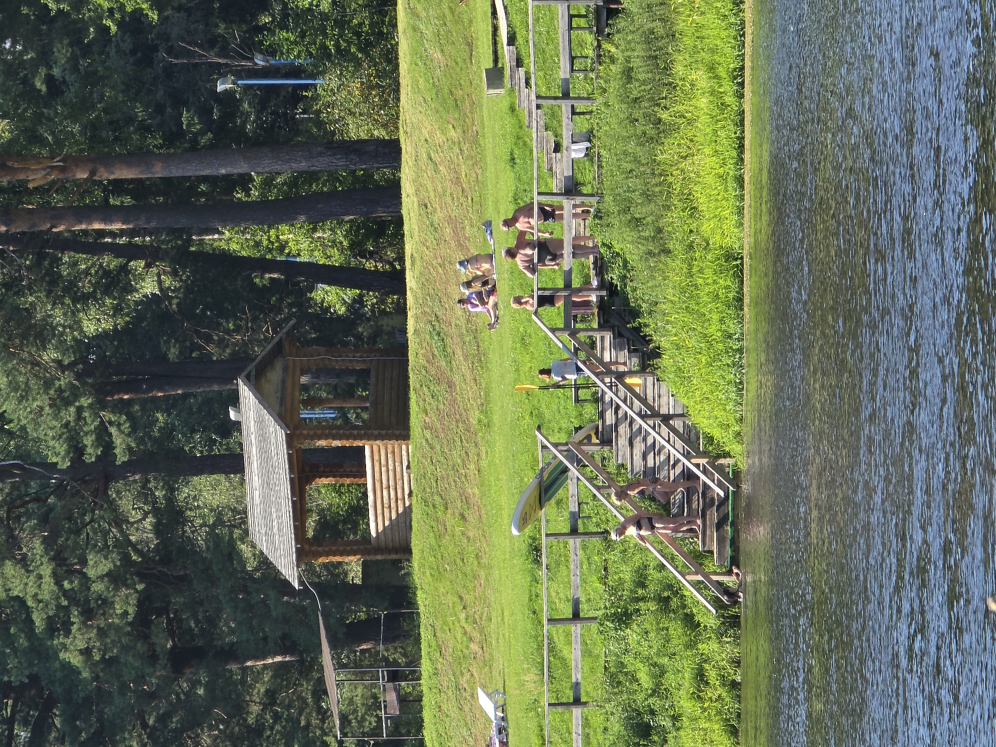 An outdoor recreational area with a wooden gazebo on a grassy embankment overlooks a body of water. In the foreground, a wooden staircase descends into the water, with several people in swimwear on the stairs and at the water's edge. A paddleboard is propped against the railing of the stairs. In the midground, other individuals are seen sunbathing on the grass. The background is dominated by tall trees. The scene is located in the city of Voinskaya Chast' Mchs, Russia.