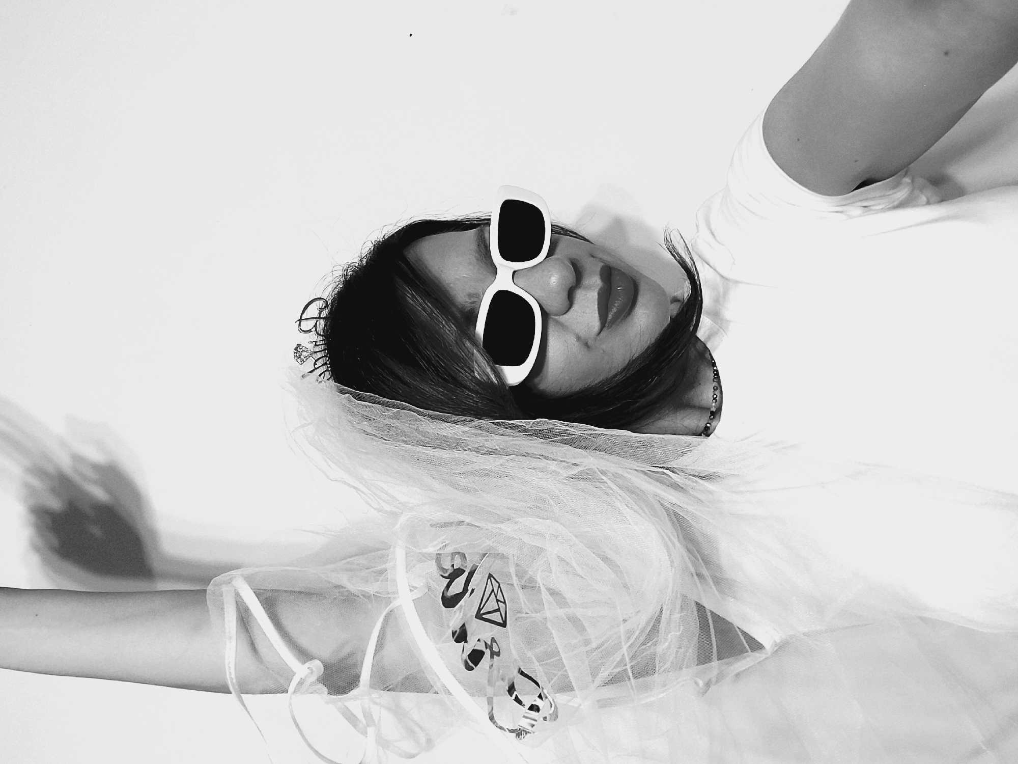 A black and white, close-up, waist-up selfie shows a person, identified as female by her attire and features, posing against a plain white wall. She is wearing a white t-shirt, white rectangular sunglasses with dark lenses, and a sparkly tiara that reads "BRIDE" on her head, partially obscured by her dark hair. A white veil is draped over her shoulder, revealing a clear plastic accessory bag with black designs, including hearts and diamonds, attached to it. Her left arm is raised, and her hand is blurred from motion. She has red lipstick on and is looking to her right with a slight smile. The lighting casts a shadow of a hand on the wall behind her left arm. There is no indication of the location being the city of Sofia, Bulgaria.