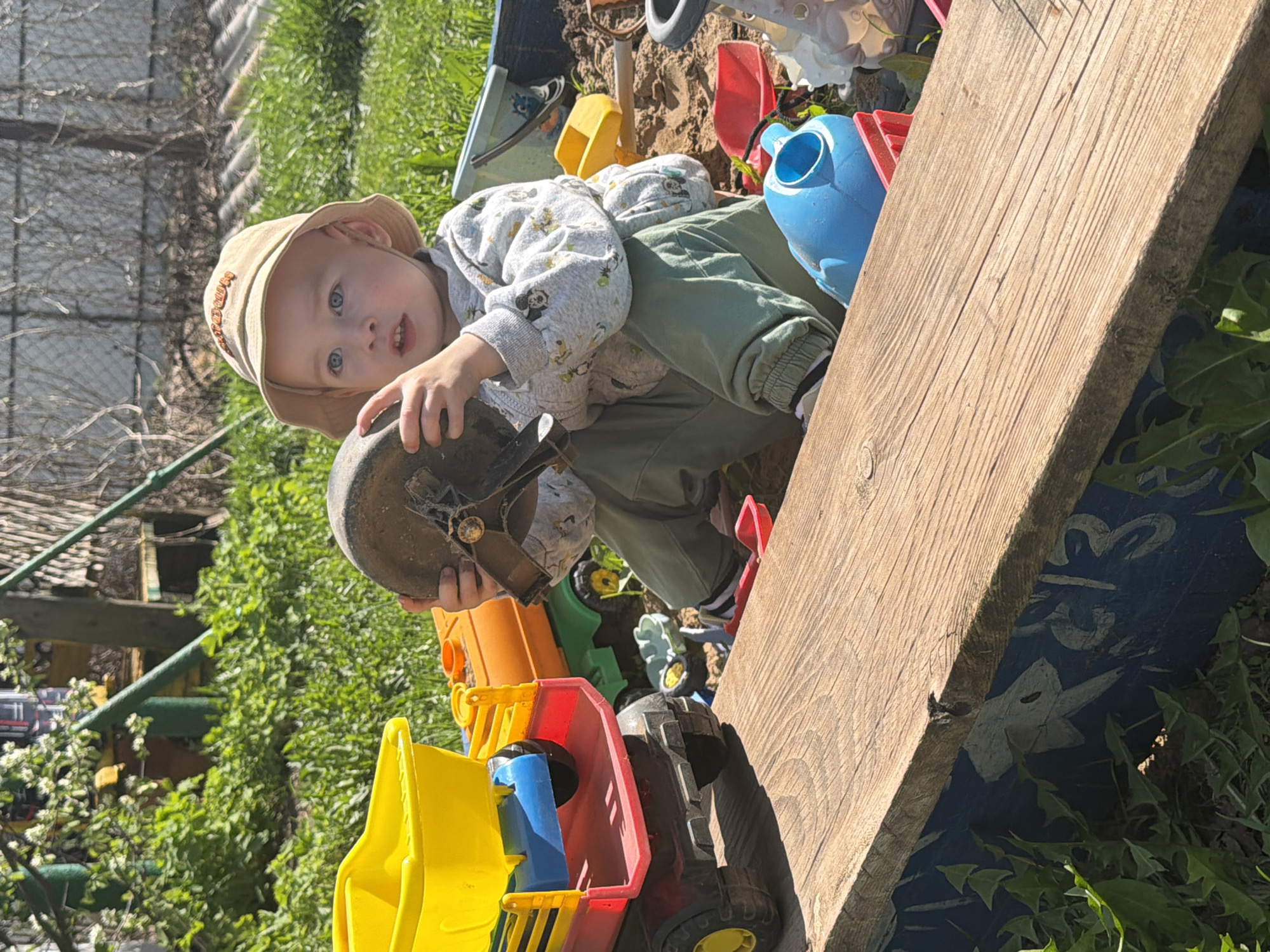 A young child wearing a light-colored hat and patterned sweatshirt sits in what appears to be a sandbox filled with various colorful plastic toys, including trucks and a watering can. The child is holding a dark-colored, rounded object, possibly part of a toy. The sandbox is bordered by a wooden plank. Grass and other vegetation are visible in the background, along with a wire fence. The location, Уральское, Russia, is a detail from the user query.