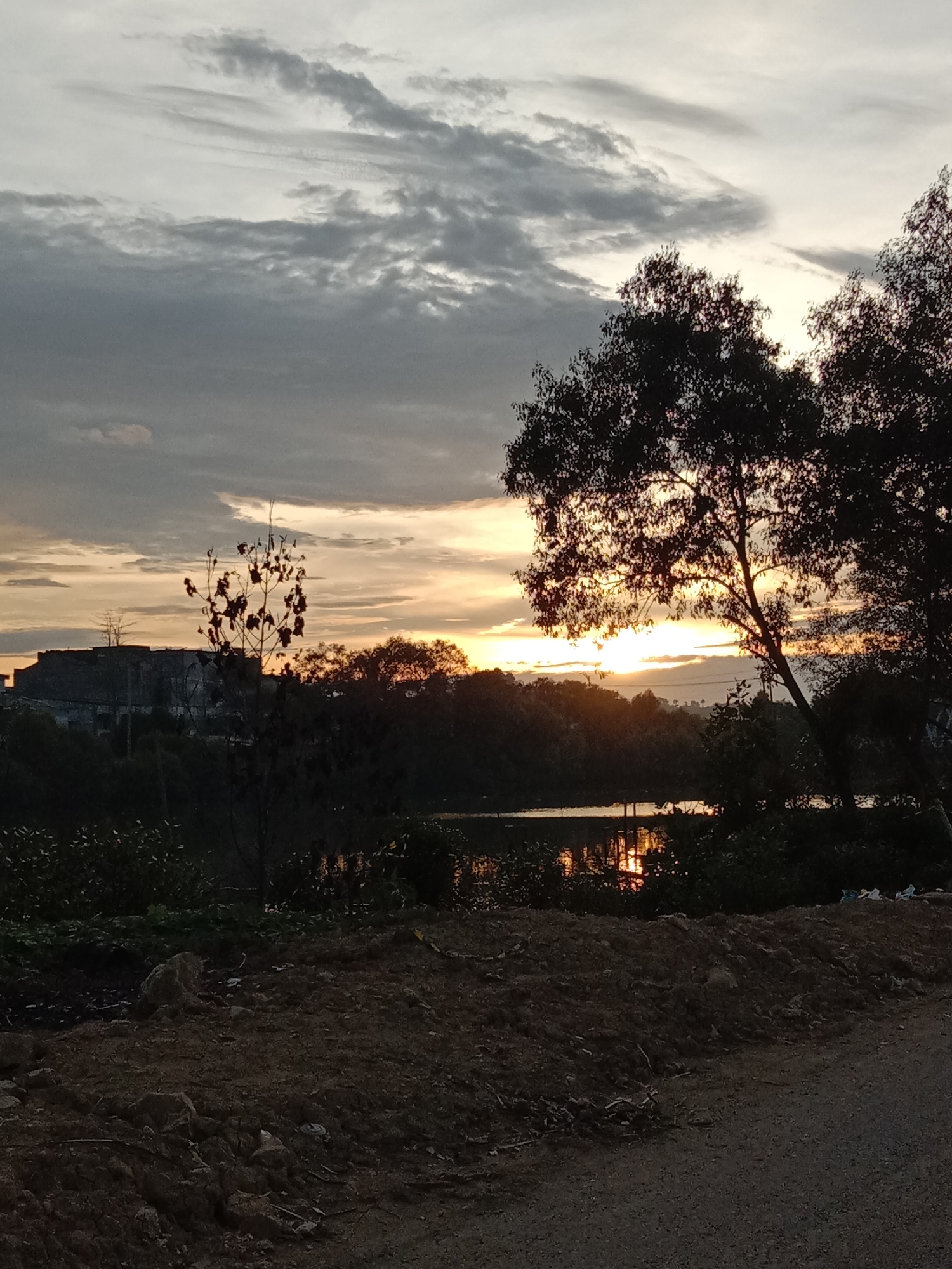 The image shows a sunset scene in Lhokseumawe, Indonesia. The sky is filled with gray clouds and a golden glow from the setting sun, which is partially obscured by trees. The sun reflects on the water surface of a small lake or pond. In the foreground, there is a dirt road or path. Vegetation, including trees and shrubs, lines the edge of the water and the path. In the distance, a building is visible on a hill.