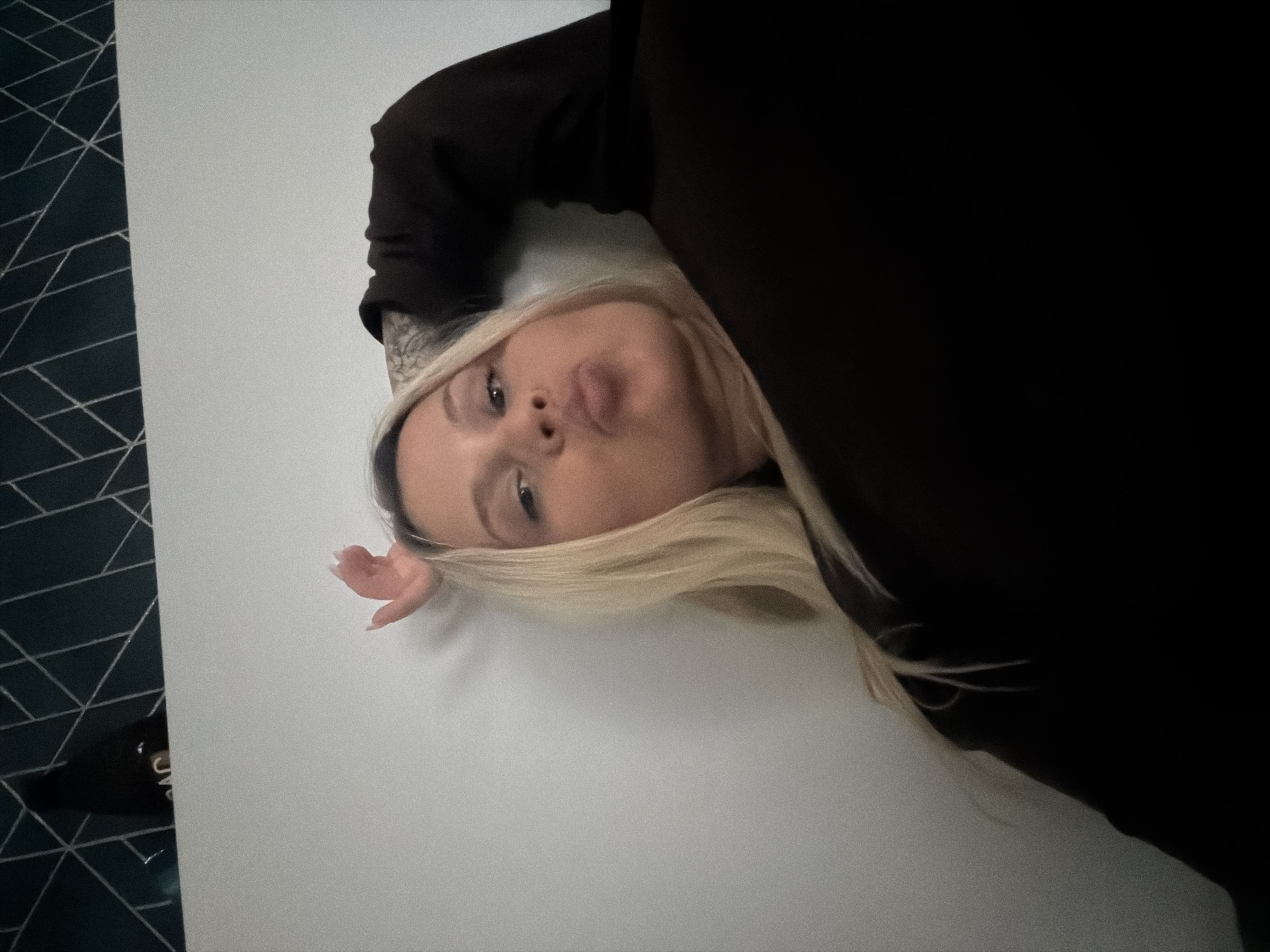 A blonde woman is lying on a white surface with a dark blue and white geometric patterned background on the left side of the frame. The woman is wearing a dark-colored top and is facing the camera with slightly pursed lips. Her right arm, adorned with a tattoo, is raised near her head. The setting's location in Sarapul, Russia, cannot be determined from the provided image.
