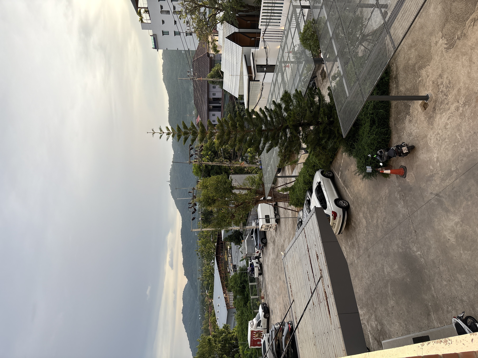 The image shows a view of a parking lot and surrounding buildings in Chalong, Thailand. The scene is dominated by a large, modern parking structure with a metal mesh roof. Several cars and motorcycles are parked in the lot. In the background, there are residential buildings, a large warehouse-like structure with the word "WARRIOR" visible, and lush green hills. The sky is overcast with some light breaking through, suggesting it might be early morning or late afternoon. A tall evergreen tree stands prominently in the foreground. There are no people actively engaged in any notable actions. The overall impression is one of a developed urban or semi-urban area with functional infrastructure.