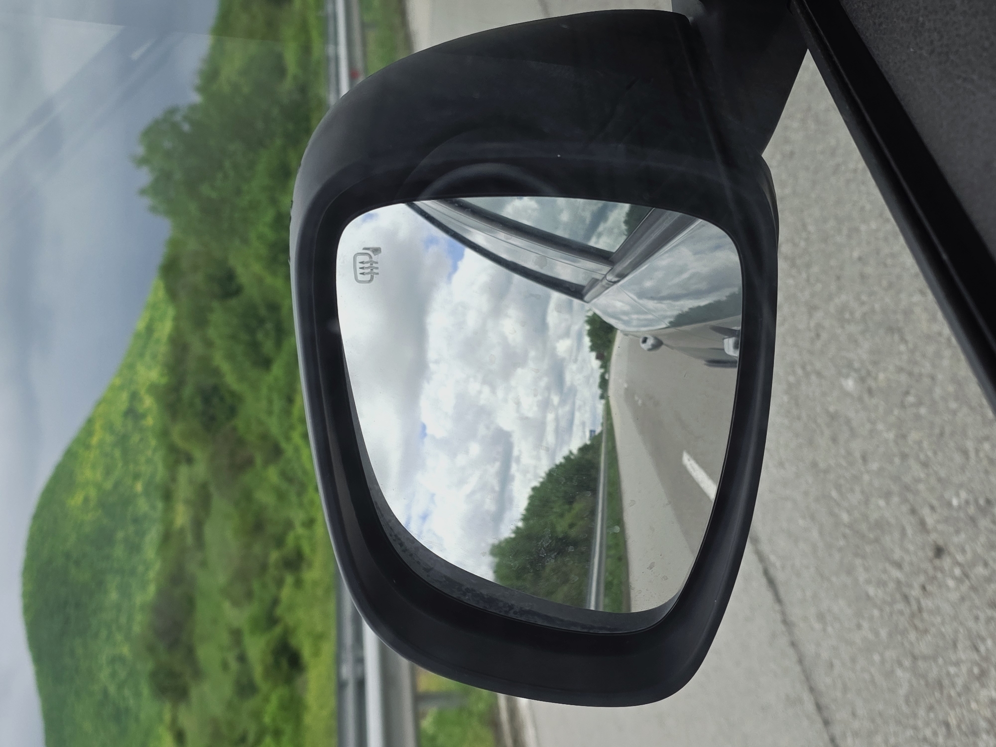 The image shows the side view mirror of a car, with the reflection showing a road, sky with clouds, and a grassy area with trees in the distance. The car appears to be driving on a highway, as suggested by the road markings. The reflection in the side mirror suggests a clear day with some clouds. The car is situated in Nevsha, Bulgaria based on the geographic location details.