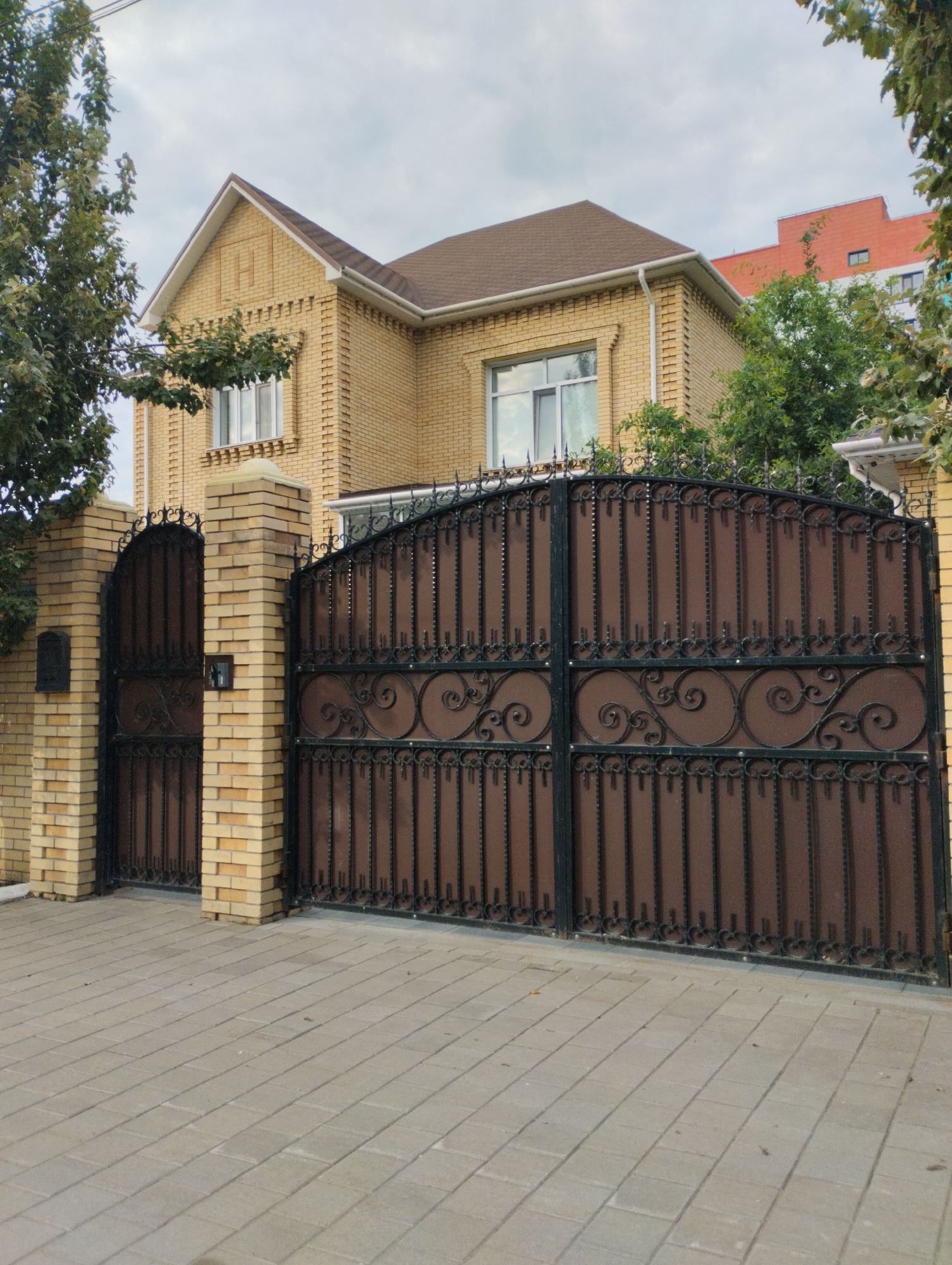 The image depicts a large, ornate wrought iron gate and a small pedestrian gate, both with a brown infill and black decorative elements, set within a brick fence. The fence is constructed of light-colored bricks and topped with a cream-colored coping. A yellow brick house with a brown roof and white trim is visible behind the gates. A large tree with green foliage is present to the left of the house, and other greenery is visible in the background. The ground in front of the gate is paved with light gray interlocking pavers. The scene appears to be a residential property in a city setting. Specifically, this is a residential property with a prominent decorative iron gate and a modern brick house. The house is a two-story structure with a light yellow brick exterior and a brown shingled roof. The gate is large and imposing, featuring intricate scrollwork and vertical iron bars. A smaller pedestrian gate is located to the left of the main gate. The fence surrounding the property is also made of brick. The overall impression is one of a well-maintained and affluent residence. The location is identified as Kostanay, Kazakhstan.