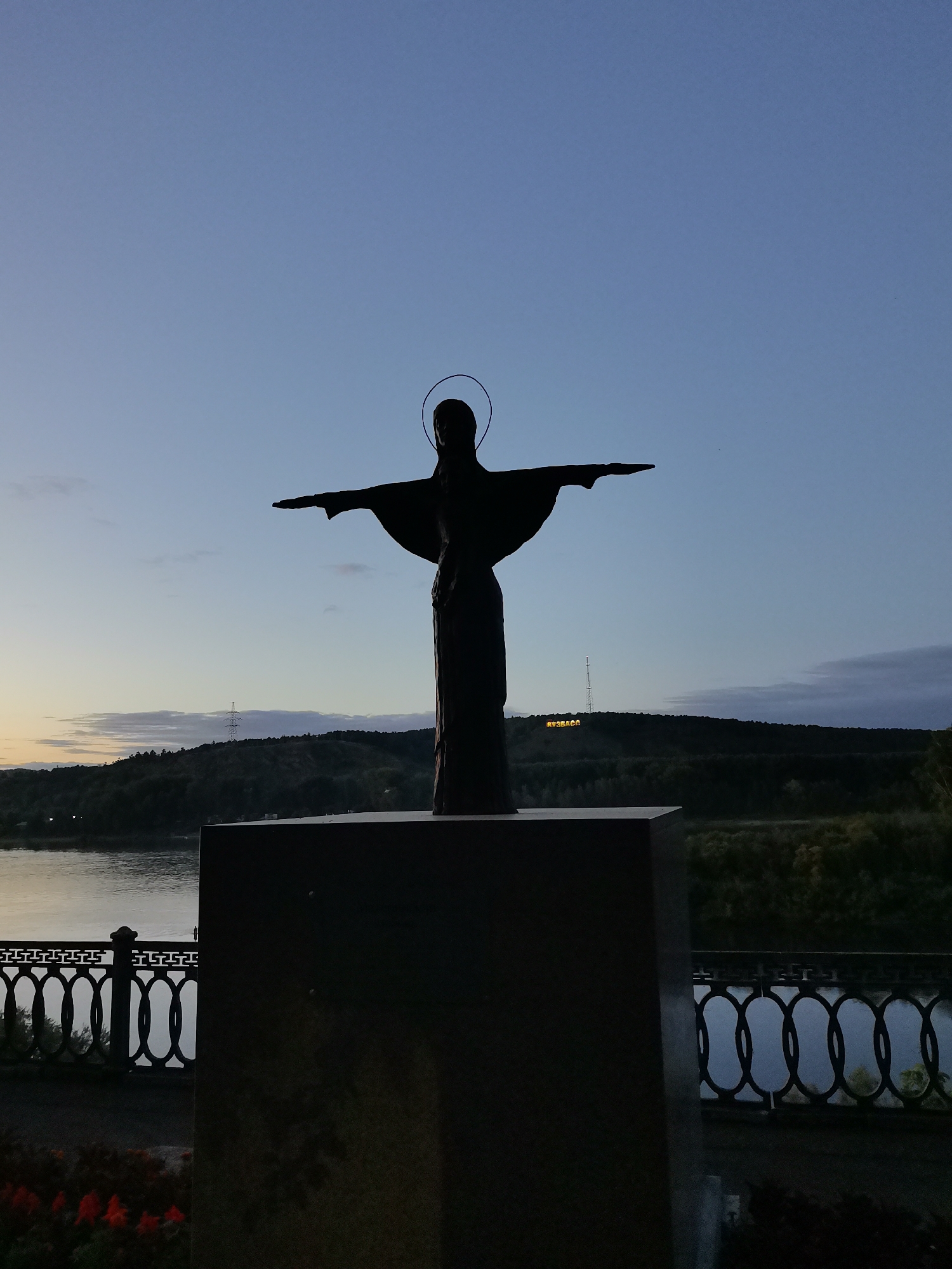 A silhouette of a statue of Jesus Christ, with arms outstretched and a halo, stands on a pedestal against a twilight sky. The statue is dark, obscuring fine details, but its form is clearly recognizable. In the background, a river or large body of water is visible, bordered by a decorative metal railing. Beyond the water, a tree-lined hill is illuminated with a sign that reads "КУЗБАСС" (KUZBASS) in yellow, illuminated letters. A tall radio tower stands on the crest of the hill. The sky is a gradient of deep blue at the top, transitioning to lighter blues and hints of orange near the horizon, indicating the end of daylight. Red flowers are visible in the foreground at the bottom left corner of the image. The overall scene suggests a memorial or viewpoint located in Kemerovo, Russia, specifically likely near the Tom River embankment, given the presence of a body of water, a prominent statue, and the regional sign.