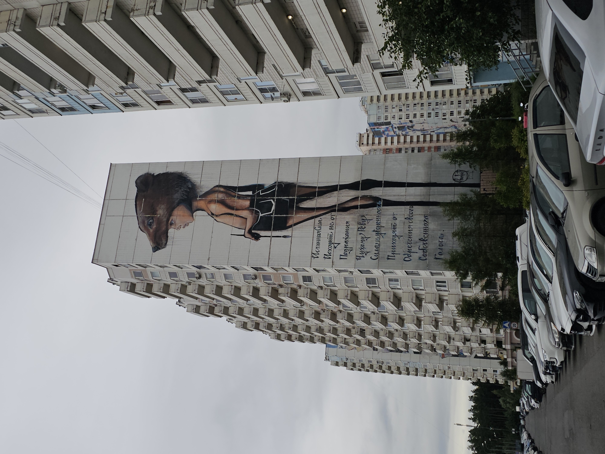 This is a ground-level, eye-level, medium shot of a tall, light-colored apartment building with a large mural painted on its side. The mural depicts a young person wearing a bear's head and holding a paintbrush. The person has slender limbs and is wearing dark shorts and a dark top. The mural is done in a realistic style with shading and detail. The building itself has numerous balconies with white railings. To the right of this building, another, similar apartment building is partially visible, also with balconies. In the foreground, a line of parked cars stretches along a paved area. The cars are various makes and models, mostly sedans. The sky is overcast and gray. The environment appears to be a residential urban area in Odintsovo, Russia, indicated by the architectural style and the presence of Cyrillic text on the mural. The trees in the background suggest a slightly suburban or park-adjacent setting.
