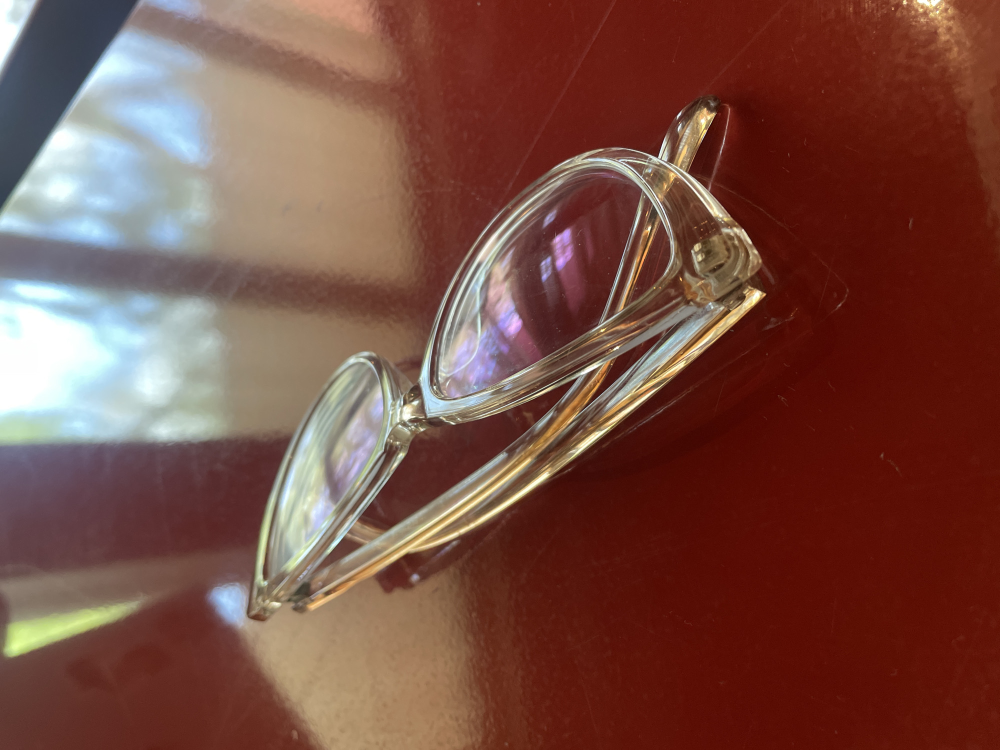 On a glossy, red-brown surface, presumably a table indoors, rests a pair of clear-framed eyeglasses. The lenses are visible and reflect light, hinting at an indoor setting. No people are present in the frame. The background is blurred and indistinct. The location in the city of Yerevan, Armenia is not discernible from the image.