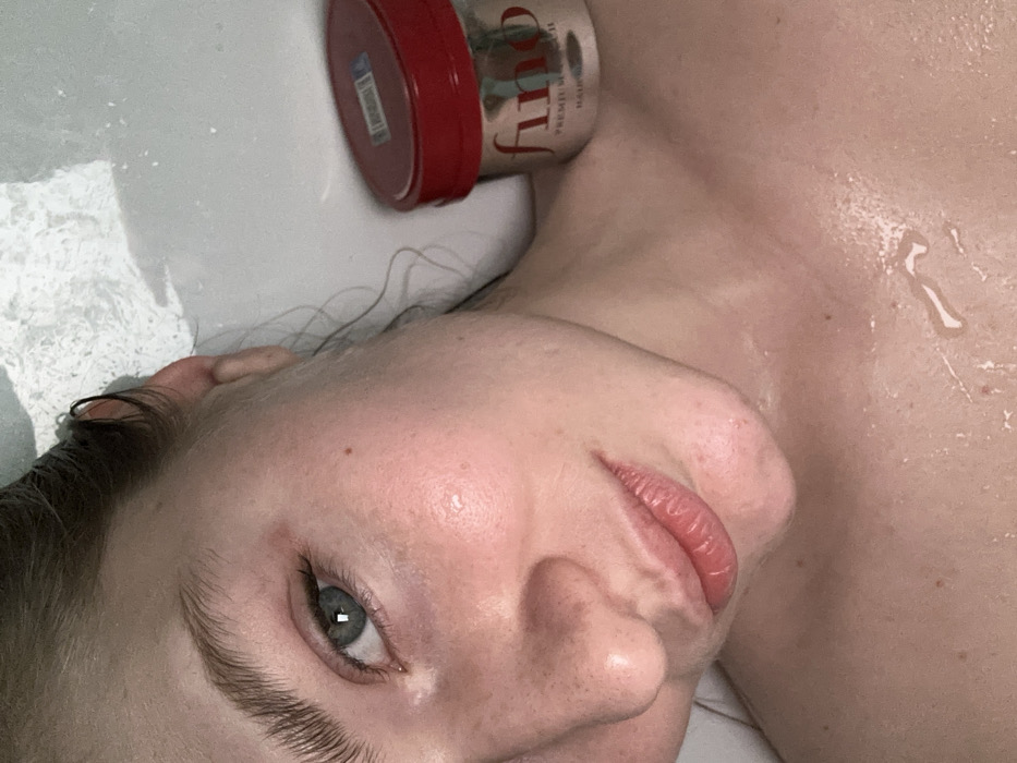 A person lies supine with their face tilted towards the camera, showing an eye with a gray iris. The face and upper chest appear to be wet. A red and silver container labeled "Fino Premium Touch Hair Mask" sits near the upper left. The person's hair is dark. A droplet of water is present on the upper chest. The background consists of what seems to be a white surface, potentially a bathtub.