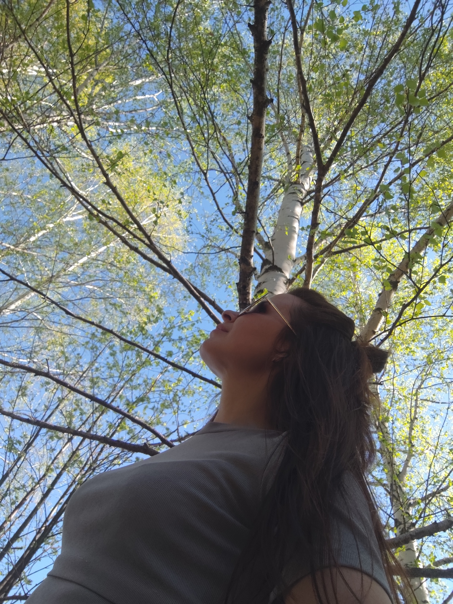 A low-angle shot depicts a woman gazing upwards at a birch tree against a clear blue sky. The woman has long brown hair and is wearing sunglasses and a gray shirt. The birch tree's white bark and thin branches extend across the frame, partially obscuring the sky. Green leaves are visible on the branches, indicating springtime. The location is Фоки, Russia.