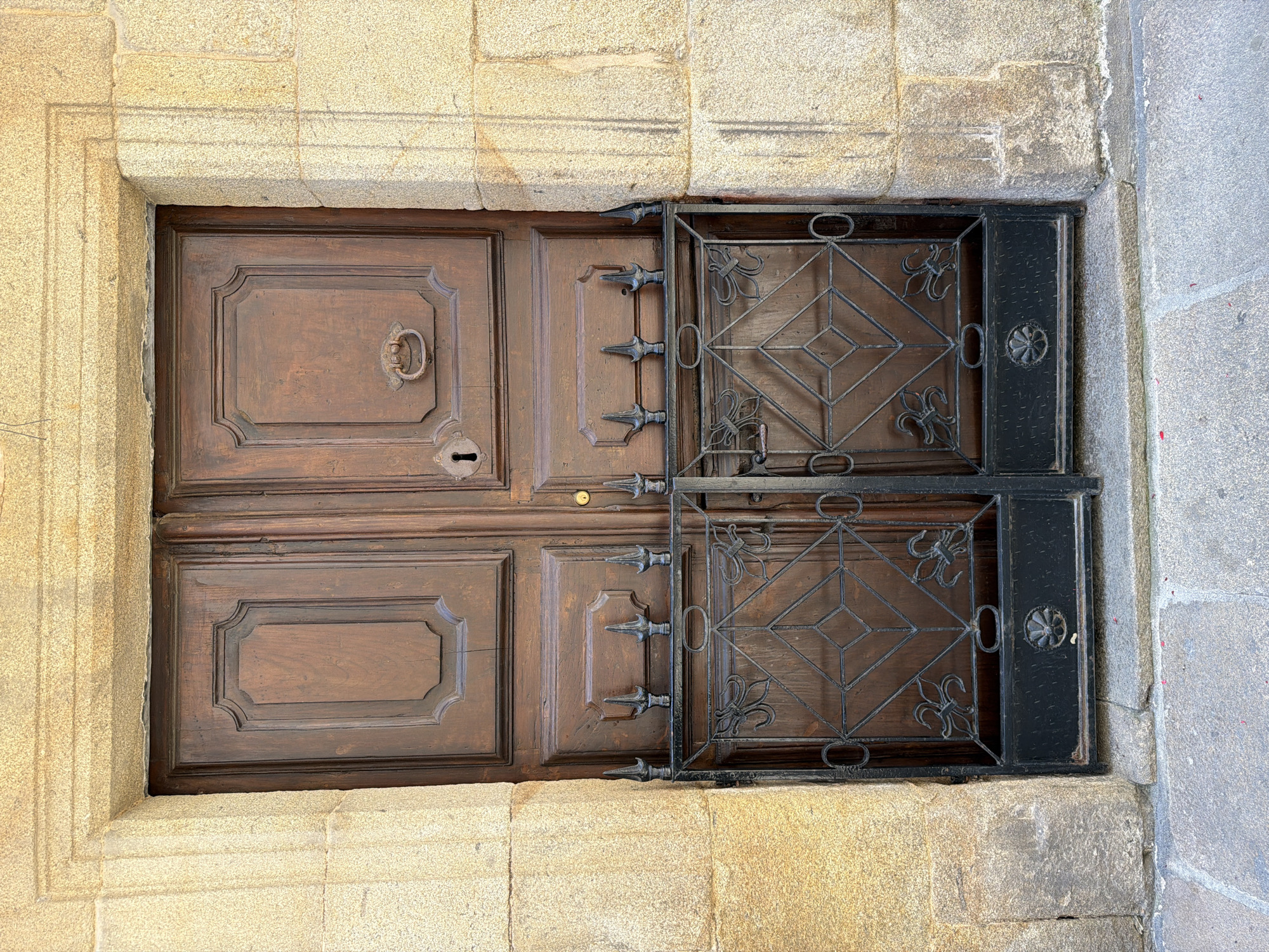 The image shows a closed, weathered wooden double door set within a stone archway in Santiago De Compostela, Spain. The doors are dark brown with carved panels and a metal door handle on the right door and a keyhole on the left. Below the doors, a decorative black metal gate with ornate scrollwork and fleur-de-lis motifs is attached. The stonework surrounding the door is rough-hewn and appears aged. The ground in front of the door is paved with large, irregular stones. The overall scene suggests an entrance to an older building, possibly a historic church or cathedral.
