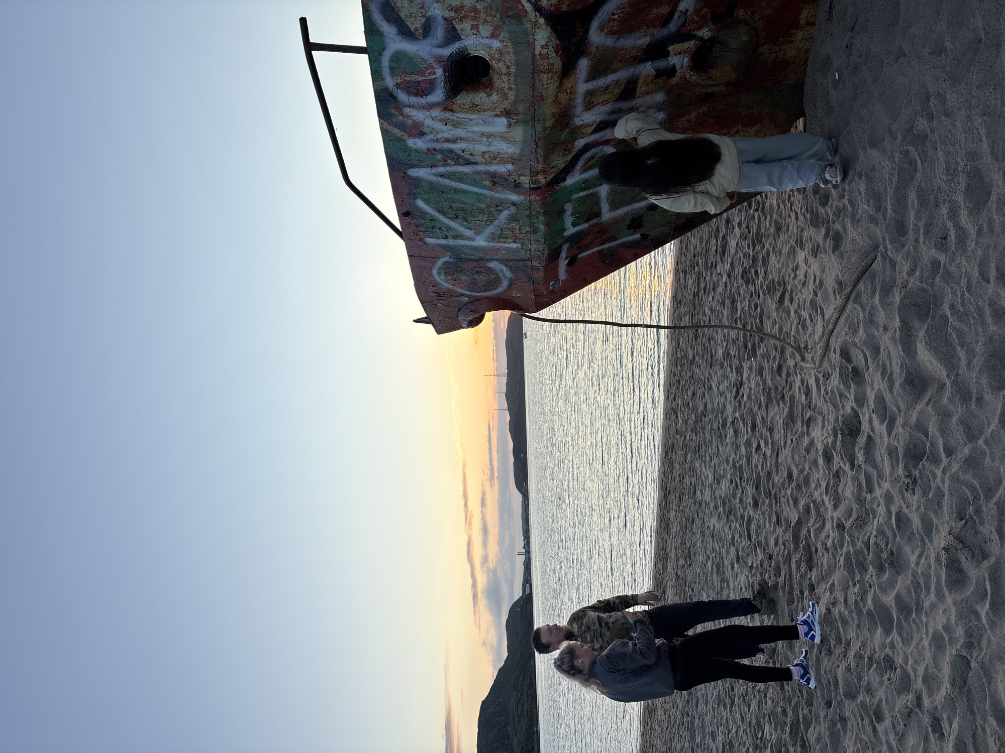 This image depicts a sunset scene on a sandy beach in Teriberka, Russia, adjacent to a body of water. In the foreground, a partially submerged shipwreck, heavily vandalized with graffiti, dominates the right side of the frame. A young girl with long dark hair, wearing a light-colored jacket and blue pants, stands with her back to the camera, touching the graffiti on the shipwreck. To the left, a man and a woman stand on the sand, looking out towards the water and distant hills. The man is wearing a camouflage jacket, and the woman is wearing a dark jacket and black leggings. The sky above is a gradient of blue and pale orange, indicating the setting sun, with clouds on the horizon. The water is calm, reflecting the sky's colors. The sandy beach is textured with ripples and footprints.