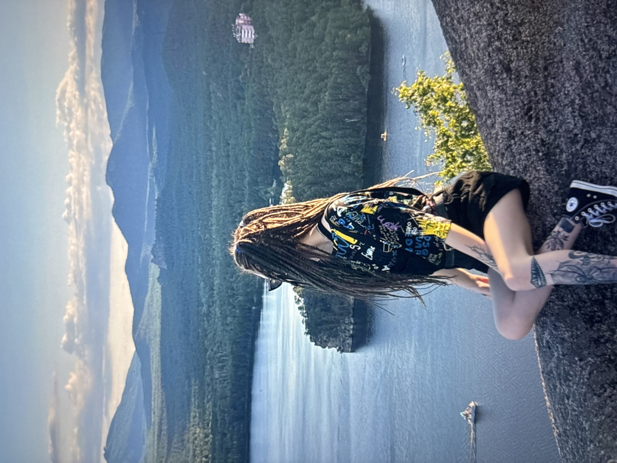 A person with dreadlocks sits on a large rock, overlooking a wide body of water surrounded by dense green trees. In the distance, rolling mountains are visible under a sky with scattered clouds. Several boats are on the water, and a building is nestled among the trees on the far bank. The person is wearing a black t-shirt with a colorful graphic print and black shorts. Their legs are crossed, and they have tattoos on their shins and calves. They are also wearing black sneakers. The lighting suggests it is late afternoon or early evening. The location is not Omsk, Russia; it appears to be a natural landscape with a lake or river.