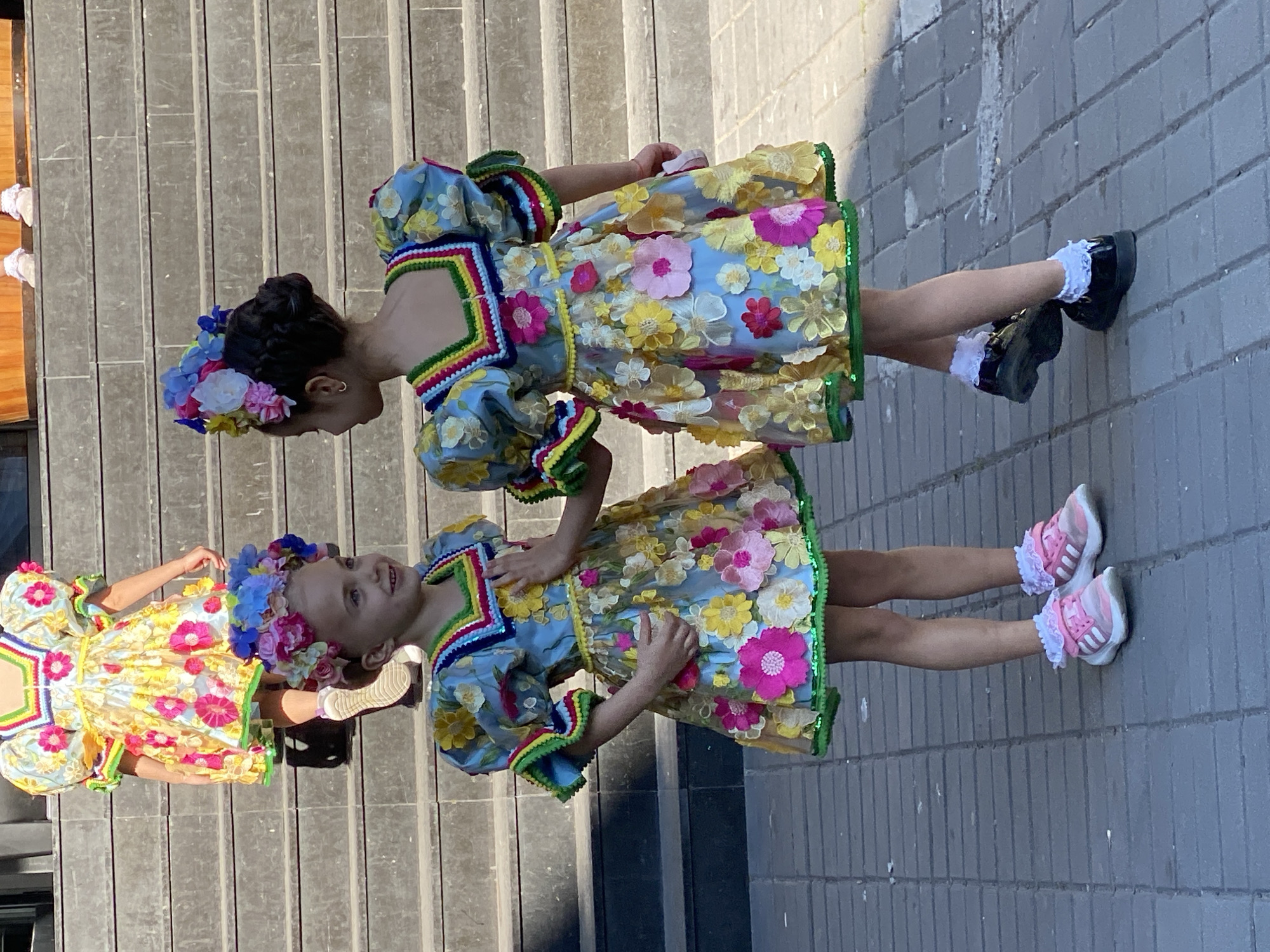 Two young girls in floral dresses and flower crowns stand on a brick surface near stone steps. One wears pink and white sneakers, while the other wears black shoes with white socks. A third figure in a similar dress is partially visible. The location is Pleven, Bulgaria.