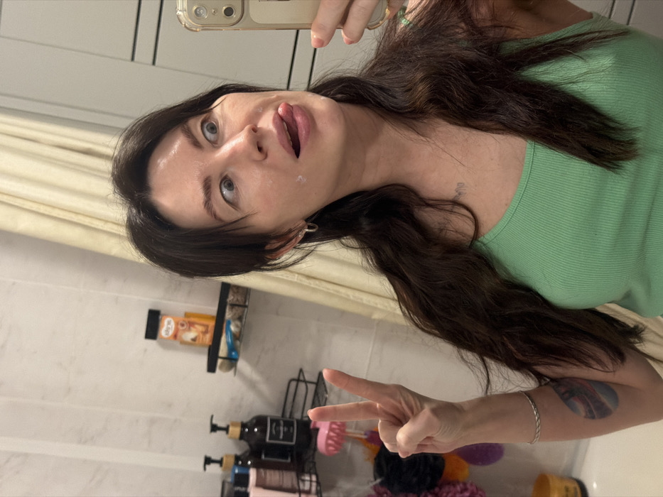 A female subject with long, dark, wavy hair and a light green ribbed tank top is depicted taking a mirror selfie in a bathroom, presumed to be located in Perm, Russia. She holds a light-colored smartphone in her right hand to capture her reflection. Her left hand is raised, displaying a peace sign. Her face exhibits a playful expression, with wide eyes directed to her right and her tongue extended slightly to her left. Small dabs of a white cream or product are visible on her left cheek and right jawline. She wears small hoop earrings. A faint, colorful, teardrop-shaped tattoo is visible on her right upper arm, and another faint, indistinct marking appears on her chest near her collarbone. The background features light-colored, rectangular wall tiles. A light-toned shower curtain is partially visible on the right. To the left, two black wire-frame shower caddies are mounted. The upper caddy holds an orange and black container labeled "Elite" in Hebrew script, alongside another smaller orange container. The lower caddy contains several pump-dispenser bottles (dark brown, light brown, clear), a pink silicone scalp massager brush, and multiple colorful bath sponges/loofahs (pink, purple, orange, black). A white bathtub rim and a yellow container are partially visible below the lower caddy. The scene is illuminated by artificial lighting.