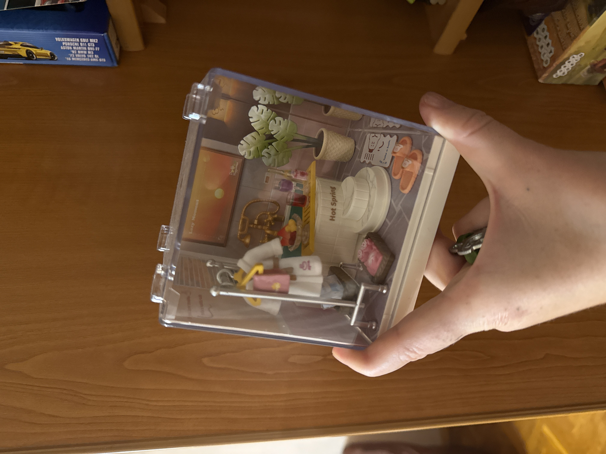 A hand holds a clear, cubed miniature diorama of a spa scene. The diorama features a robe hanging on a rack, a hot spring bath, two orange flip-flops, a potted plant, and a vintage telephone. The background of the diorama displays artwork and what appears to be a bathtub. The diorama is positioned in front of a wooden surface.