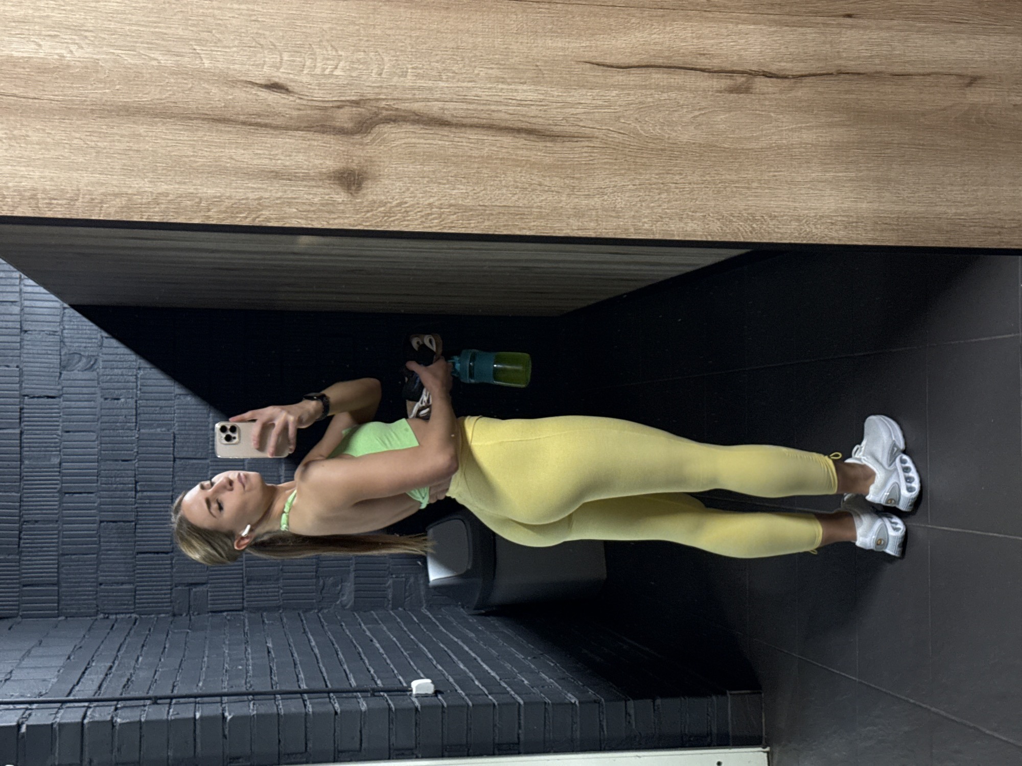 A woman takes a selfie in what appears to be a gym locker room. She is wearing a light green sports bra and yellow leggings. The background consists of a wood-paneled wall above a dark-colored, possibly black, wall with a horizontal brick-like pattern. The floor is dark. She is holding a smartphone and a water bottle. It is impossible to determine whether the location is Saint Petersburg, Russia without additional information.