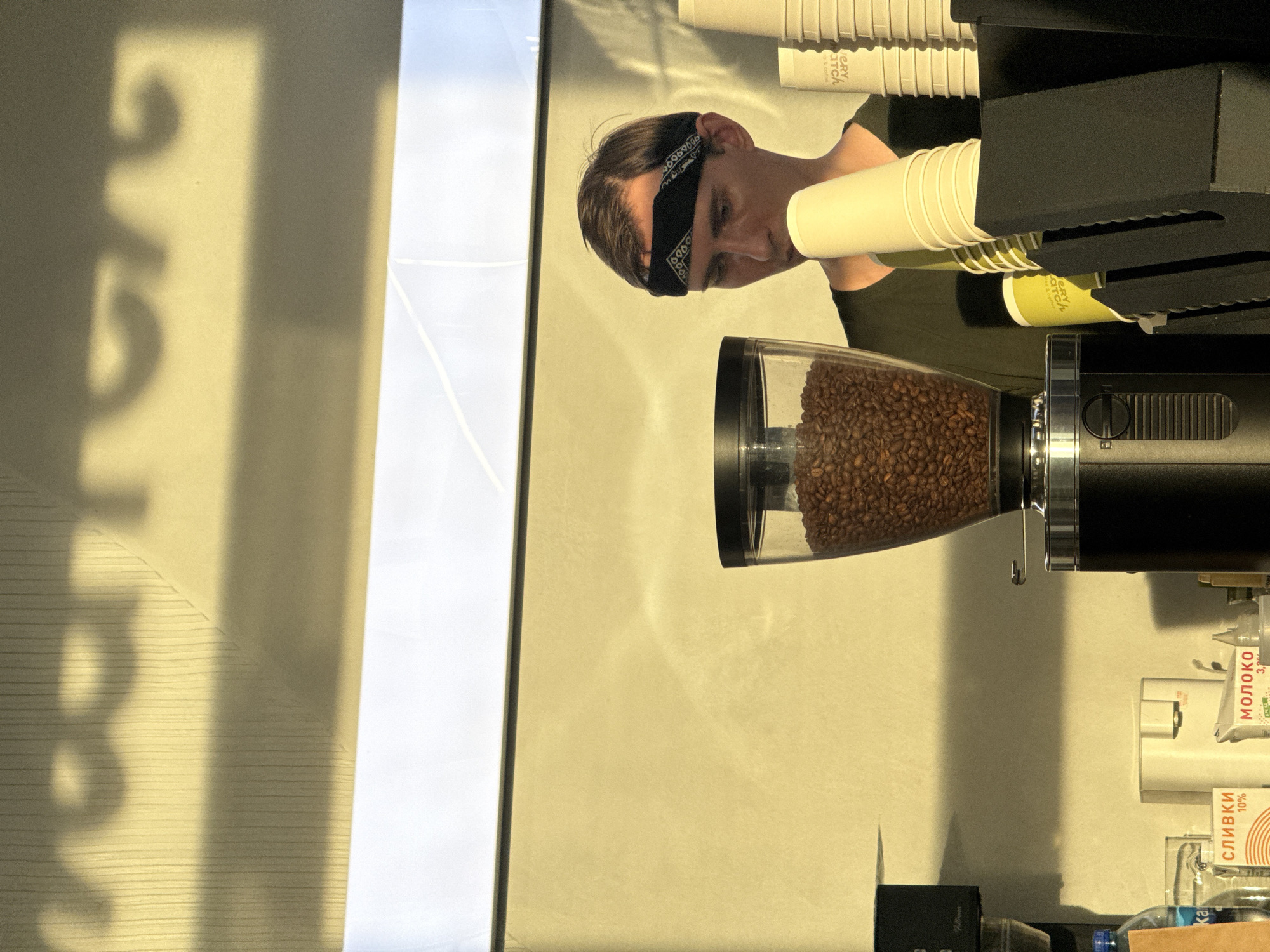 The image depicts a scene likely within a cafe or coffee shop in Nizhny Novgorod, Russia, given the context suggested by the provided Cyrillic text on some of the products. In the foreground, a coffee grinder is prominently displayed, filled with coffee beans. To the right, a stack of paper cups is visible, some of which are in a dispenser. A person wearing a dark bandana and a dark green t-shirt is partially visible in the background, appearing to be engaged in work behind the counter. The lighting suggests it is daytime, with shadows cast on the wall behind the subjects, possibly from an external source like sunlight filtering through a window. The overall environment appears to be a service area, characterized by the presence of coffee-making equipment and disposable cups.