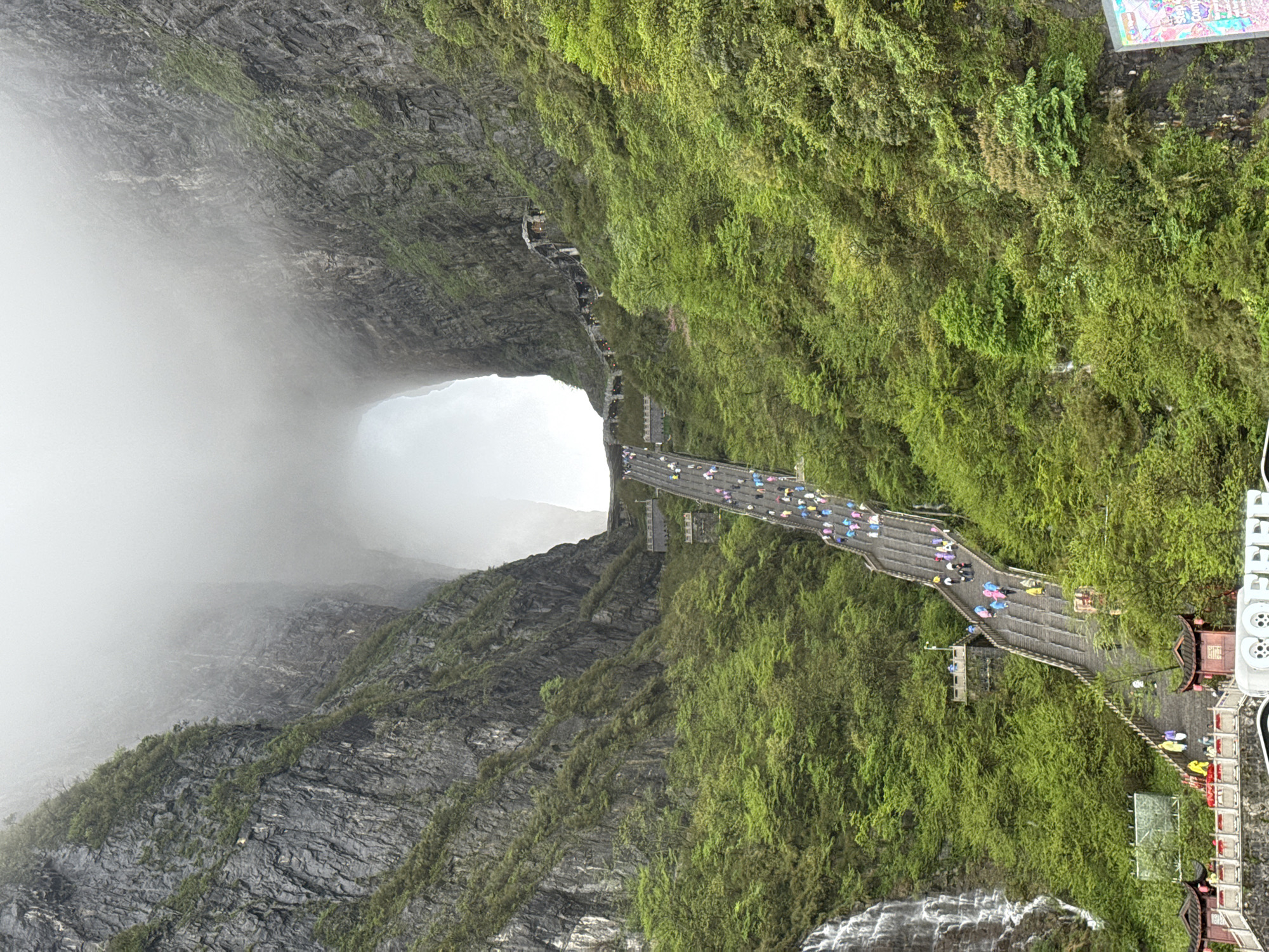 The image depicts the Tianmen Cave, also known as "Heaven's Gate," in the Tianmen Mountain National Park, located in Zhangjiajie, China. A long, stone stairway leads up to the natural archway, which is shrouded in mist and clouds. Numerous people with umbrellas are walking up the stairs toward the cave. The surrounding mountains are covered with lush green vegetation on one side and steep gray cliffs with sparse vegetation on the other. At the base of the staircase, there is a building complex with architectural details. A waterfall is visible in the lower portion of the scene.