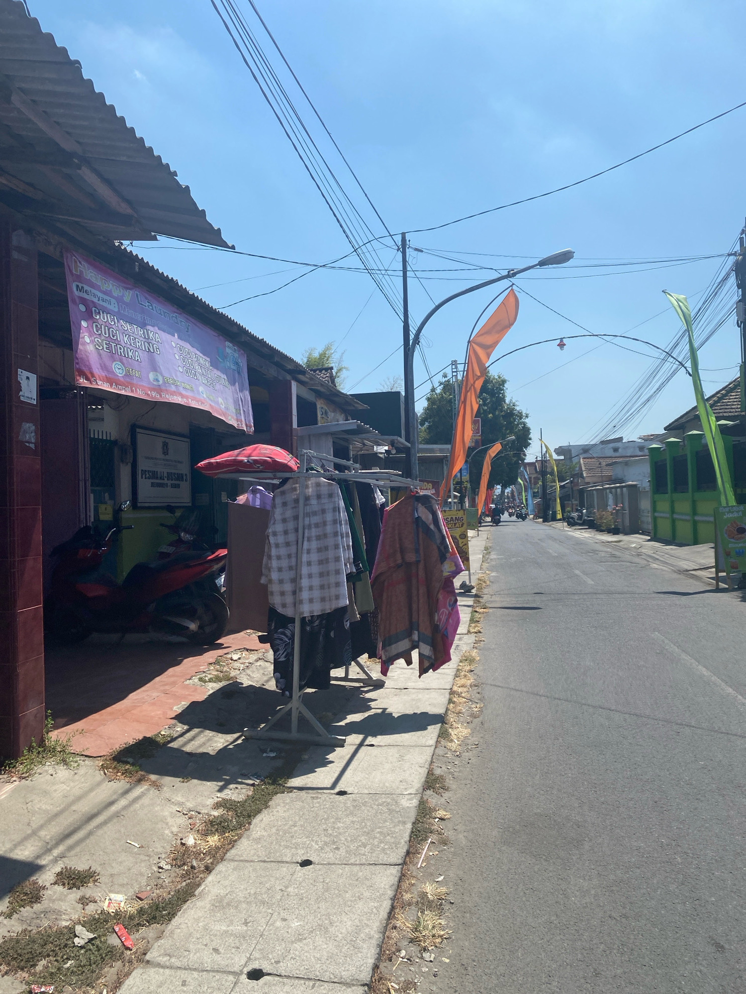 The image depicts a street scene in the city of Kediri, Indonesia. On the left, a building with a weathered corrugated metal roof houses a business with a banner advertising "Happy Laundry" services, listing "CUCI SETRIKA" (wash and iron) and "CUCI KERING" (dry cleaning). A red motorcycle is parked in front of the building. In front of the laundry business, clothes are hanging on a rack near the sidewalk. The sidewalk is made of concrete slabs and has some vegetation and debris. A red umbrella is open and positioned above the clothes rack.
The street itself is paved and appears to be a main thoroughfare, with several vehicles, including motorcycles, visible in the distance. The street is lined with buildings on both sides. On the right side of the street, there are more businesses, some with green facades, and other structures. Numerous power lines and utility poles crisscross the sky, along with decorative orange and green flags strung across the street, suggesting a festive occasion or local event. The sky is clear and blue. The overall impression is a busy, everyday street in an Indonesian city.