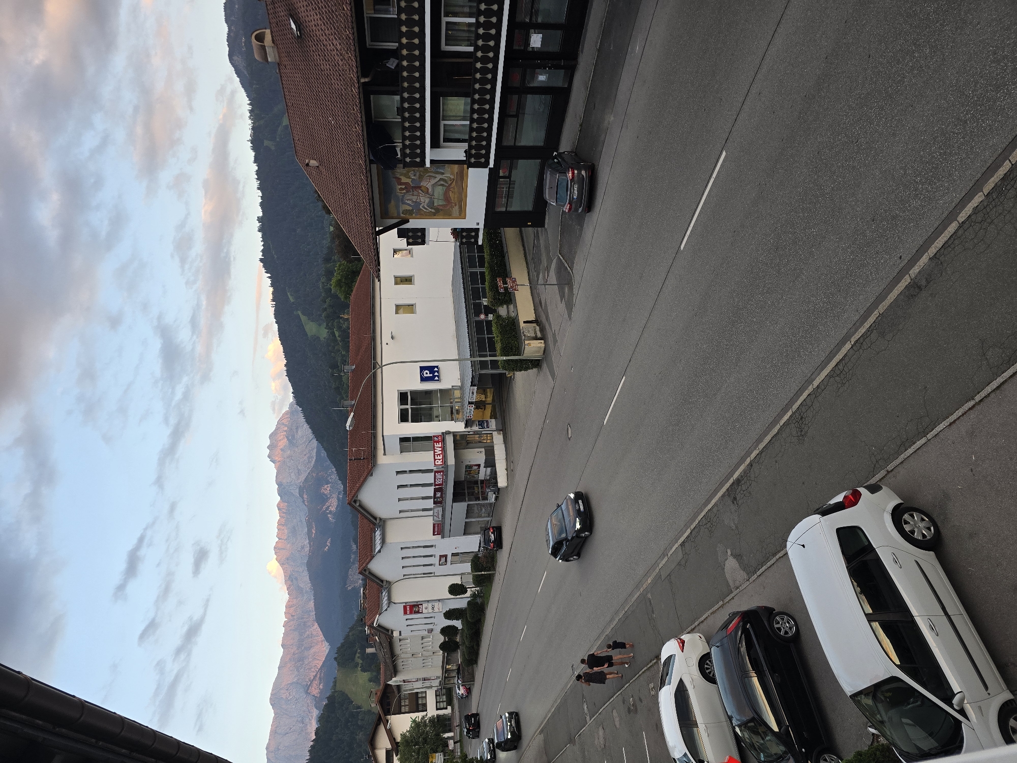 This is a medium-distance, eye-level shot of a street in Garmisch-Partenkirchen, Germany, featuring a row of buildings on the right and mountains in the background. The street is paved and has lane markings. Several cars are parked along the right side of the street and are driving on it. On the left side of the street, there are several buildings, including a white building with "REWE" signage and a building with a mural on its facade. A family of three, consisting of two adults and one child, is walking on the sidewalk on the left side of the street, facing away from the camera. The sky is partly cloudy, with the sun setting and casting a warm glow on the mountains. The overall scene depicts a typical European street scene with a blend of urban and natural elements.